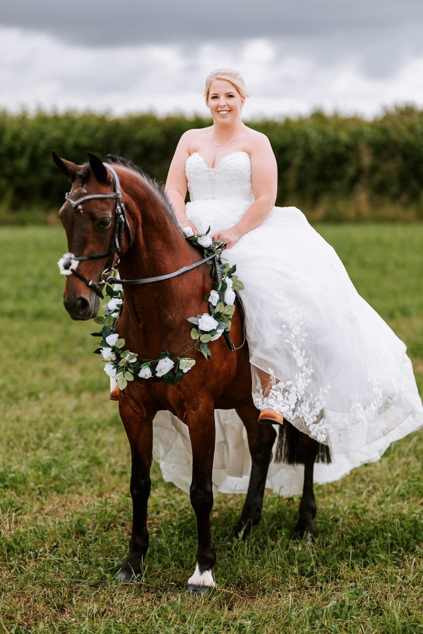 White wedding dress draped elegantly over a brown horse adorned with floral garland in a green field.