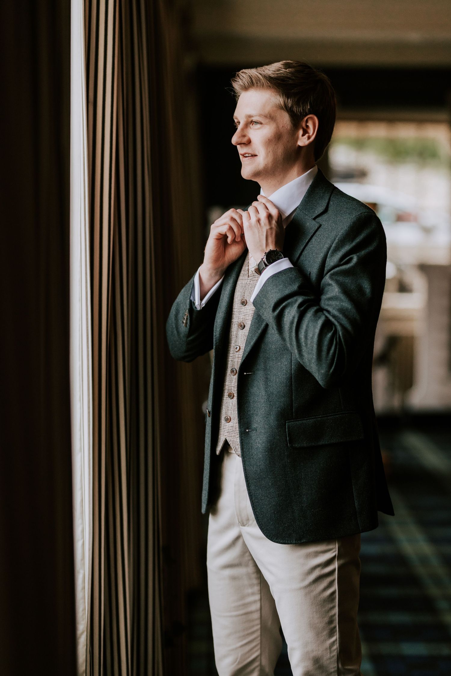 Groom in dark suit and light waistcoat looks contemplatively out window during wedding preparations.