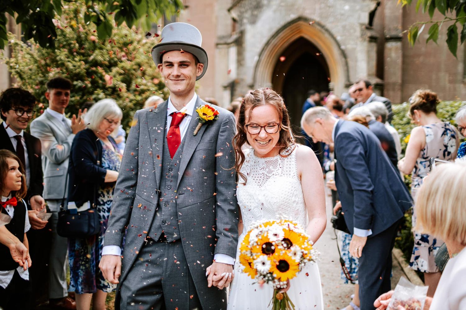 Couple exits church ceremony while holding sunflower bouquet among celebrating guests.