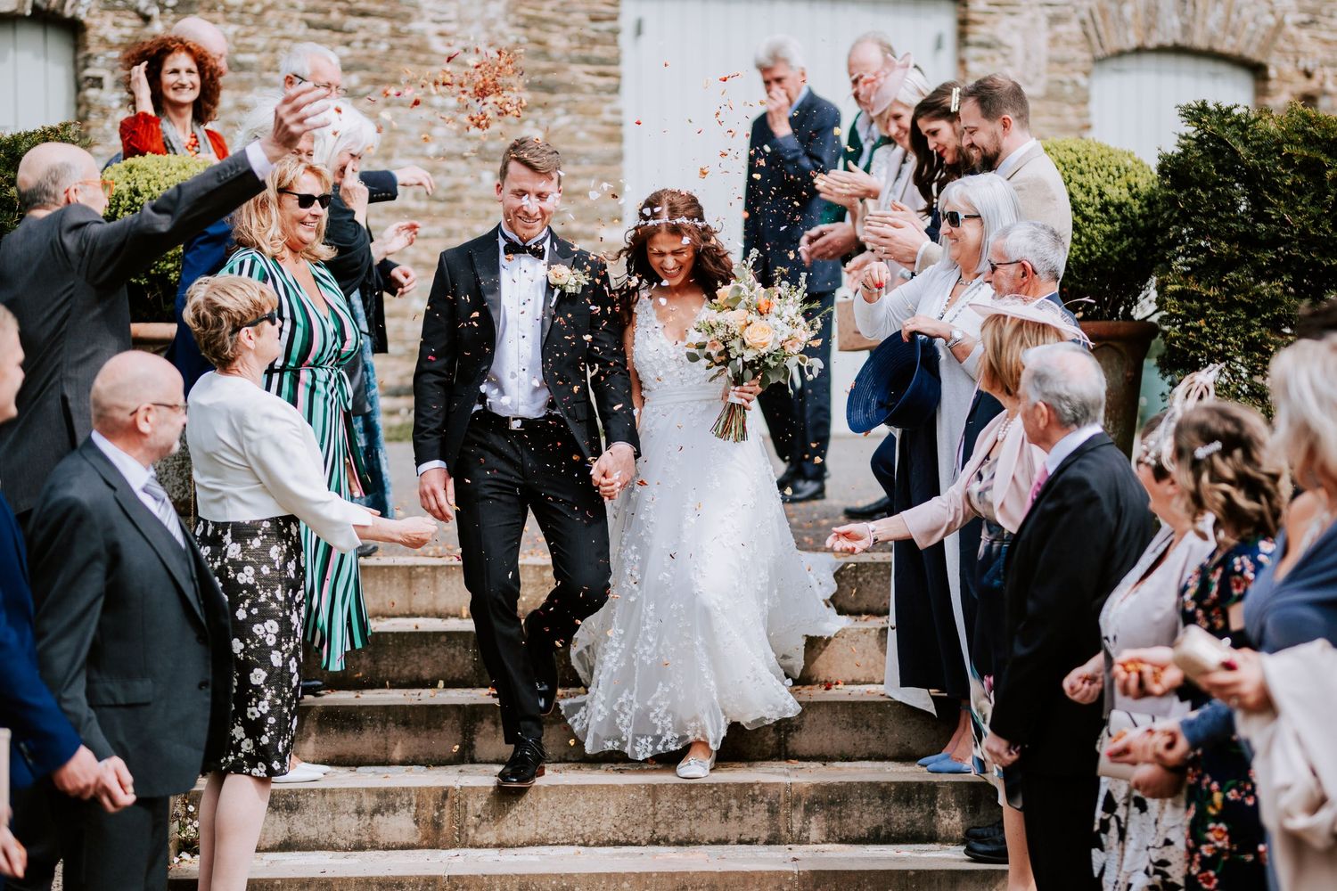 Wedding party descends stone steps while guests shower them with confetti.