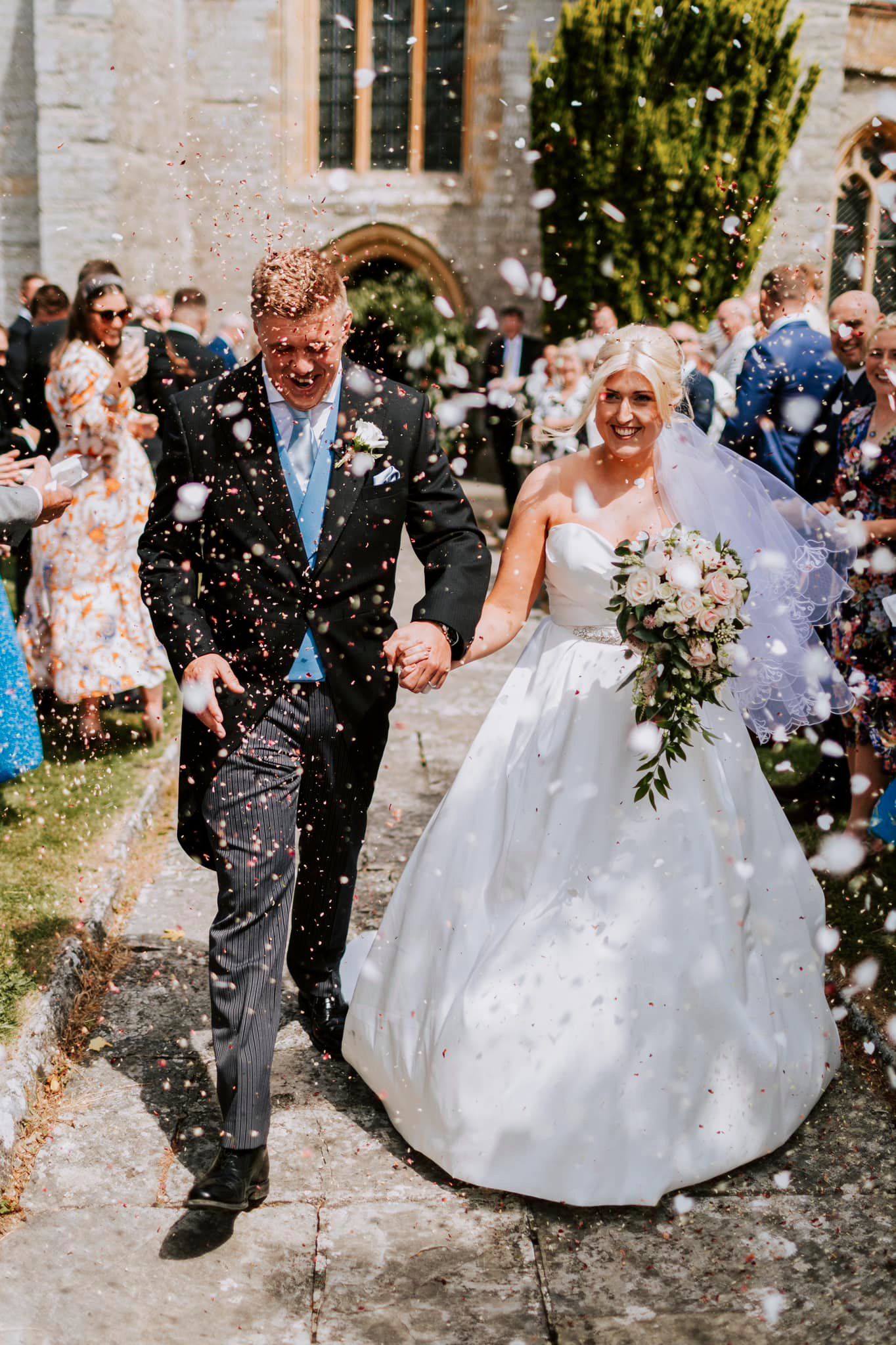 Newlyweds exit church amid shower of confetti on sunny day.