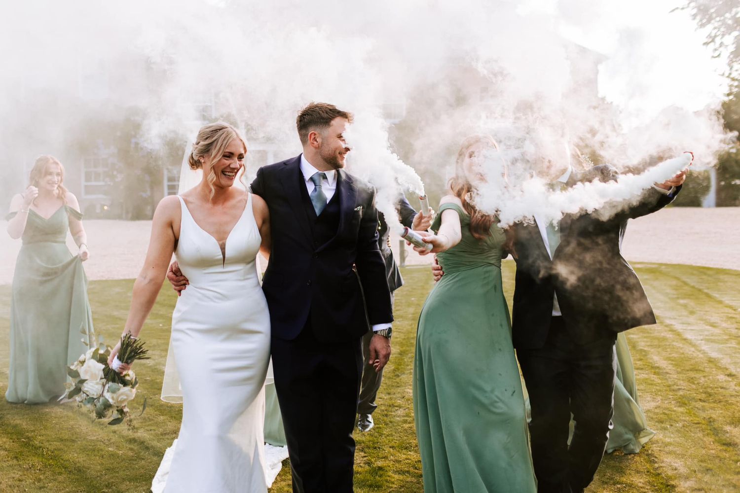 Wedding party celebrating with smoke bombs on grassy lawn.