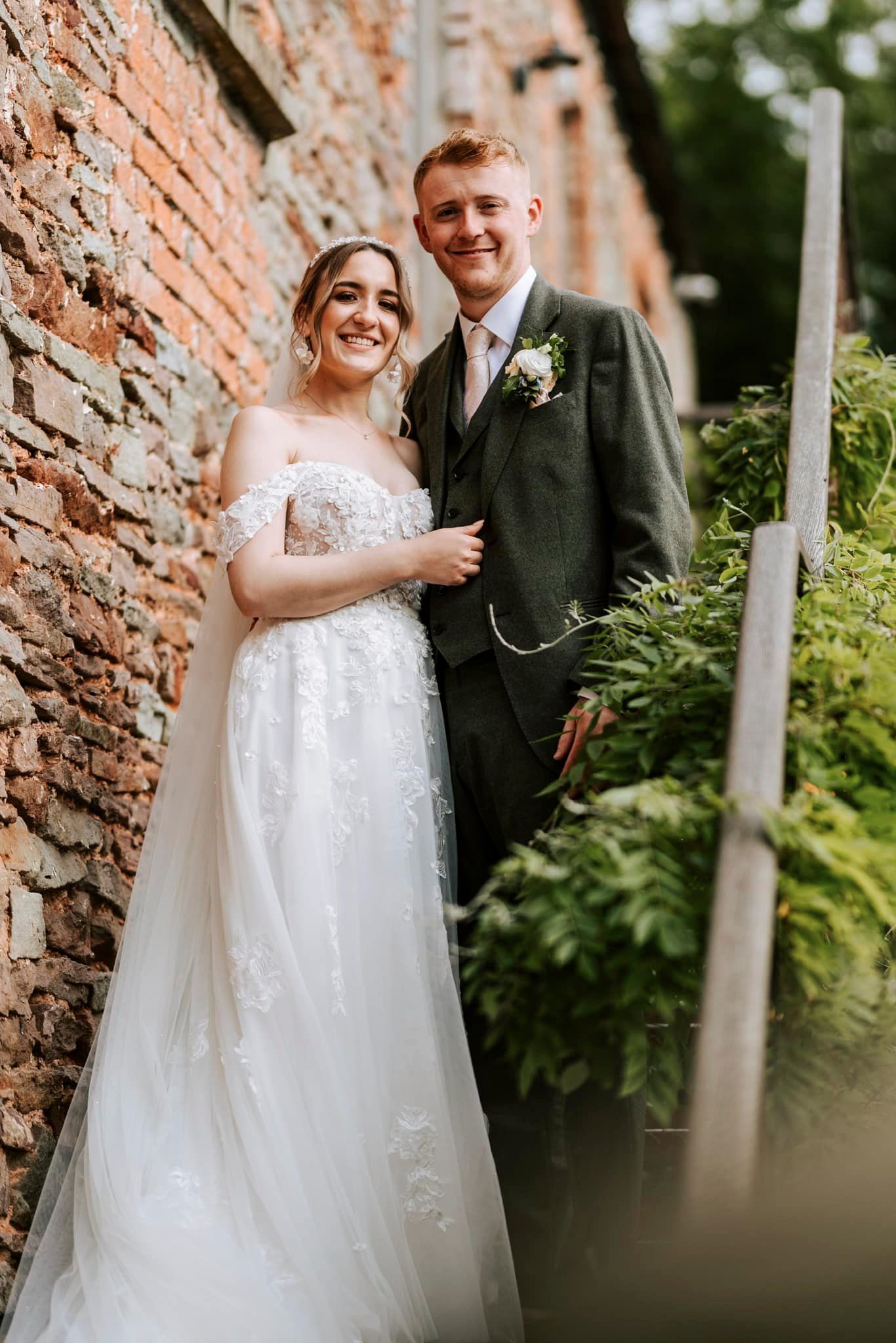 Elegant couple poses against rustic brick wall with lush greenery.