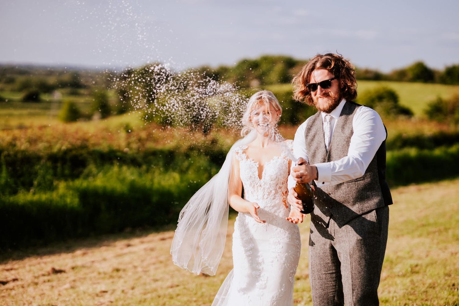 Wedding couple sprays champagne in sunny countryside celebration.