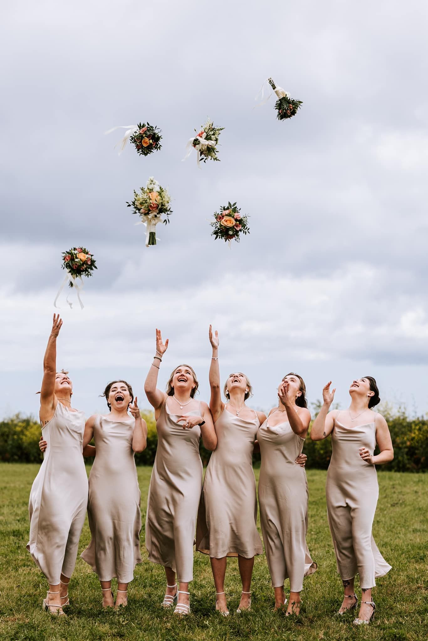 Bridesmaids in beige dresses toss bouquets in air during outdoor photo shoot.