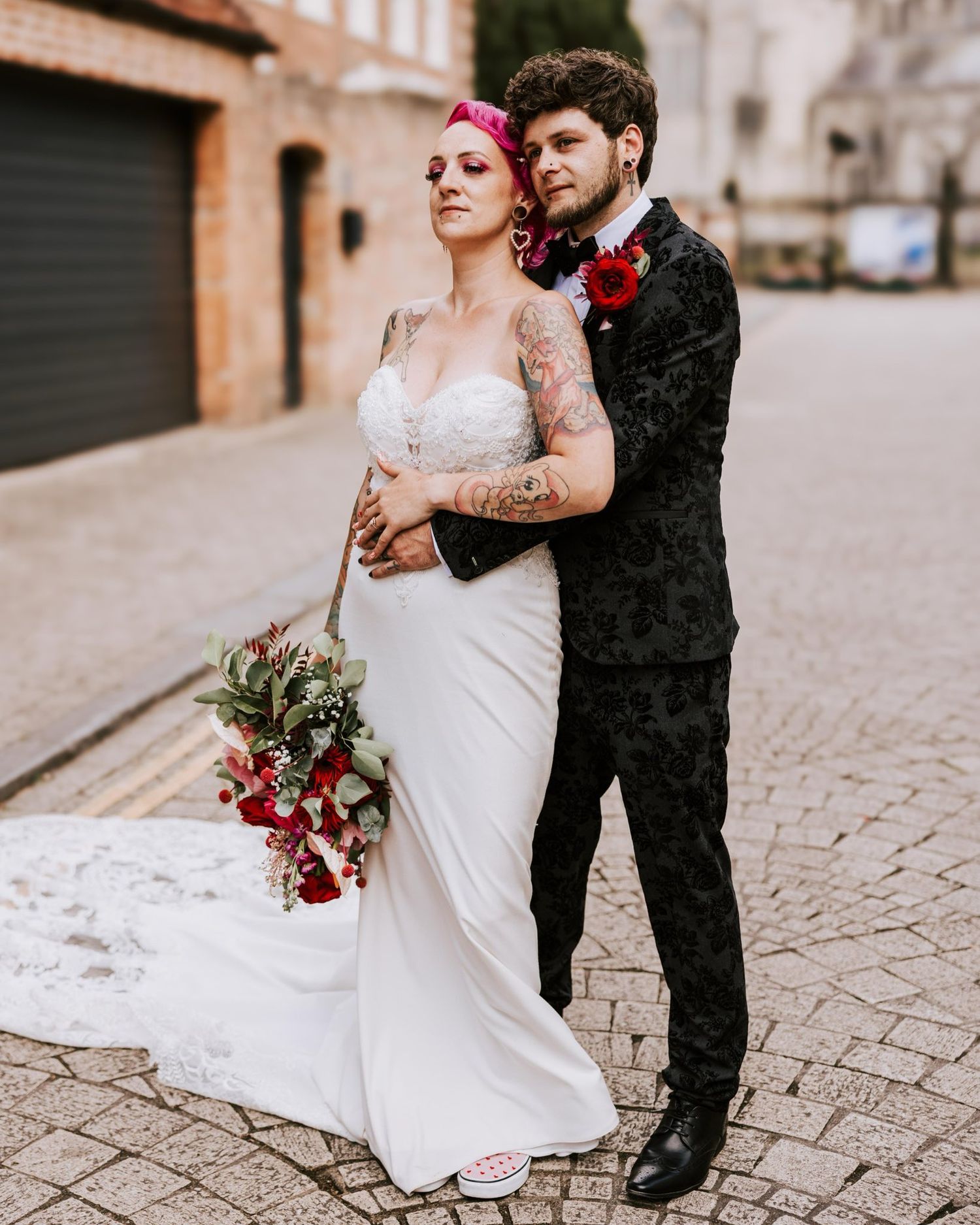 Alternative bride in white lace gown and pink hair poses with partner on cobblestone street holding red bouquet.