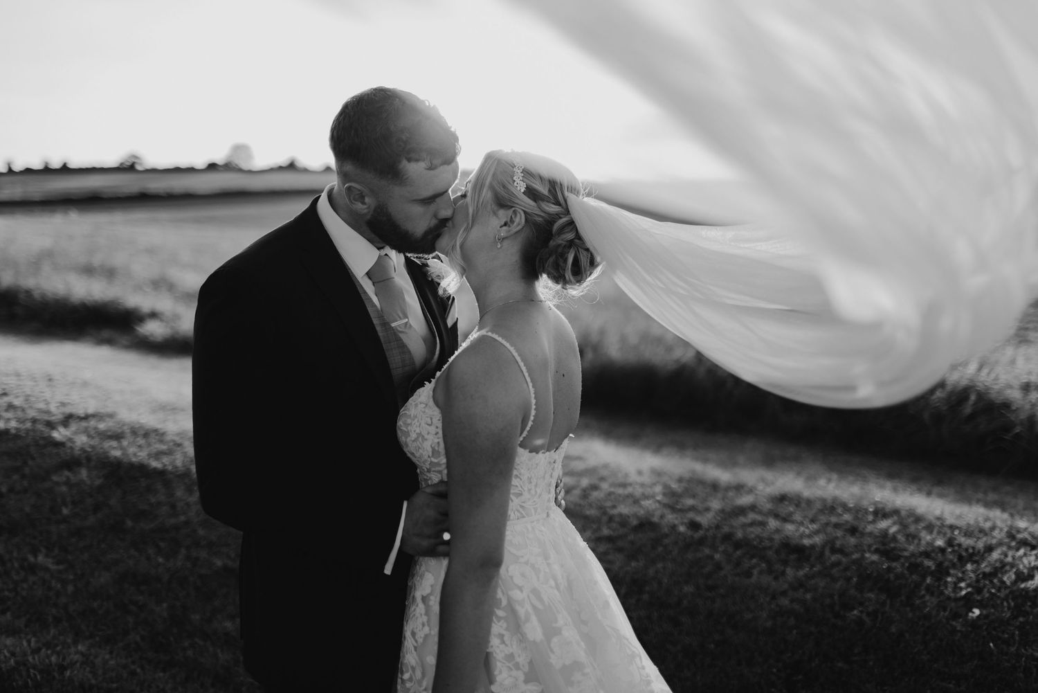 An intimate black and white wedding portrait with a flowing veil against a rural backdrop.