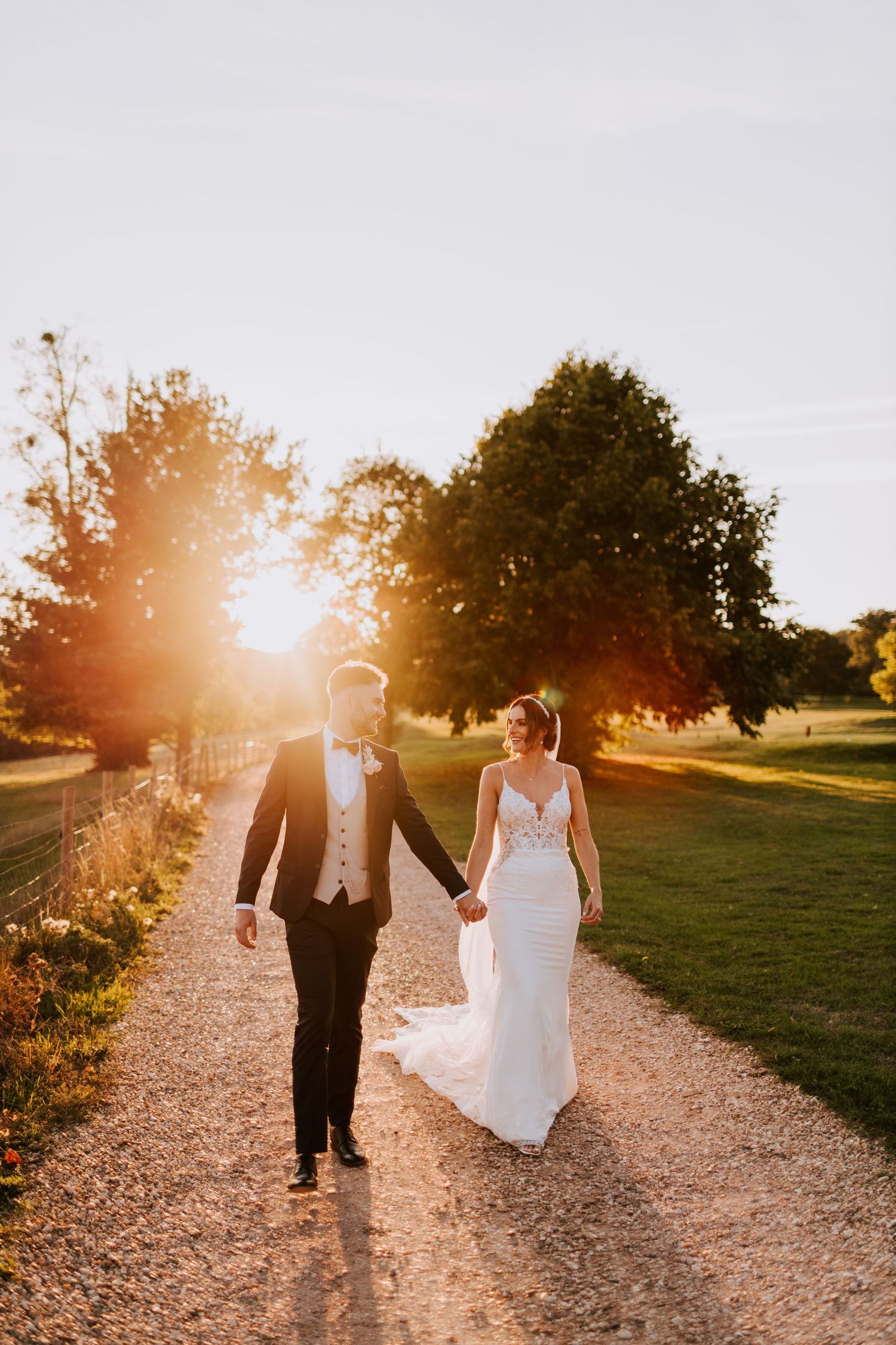 Wedding couple walks along a sunlit path during golden hour with dramatic lens flare.