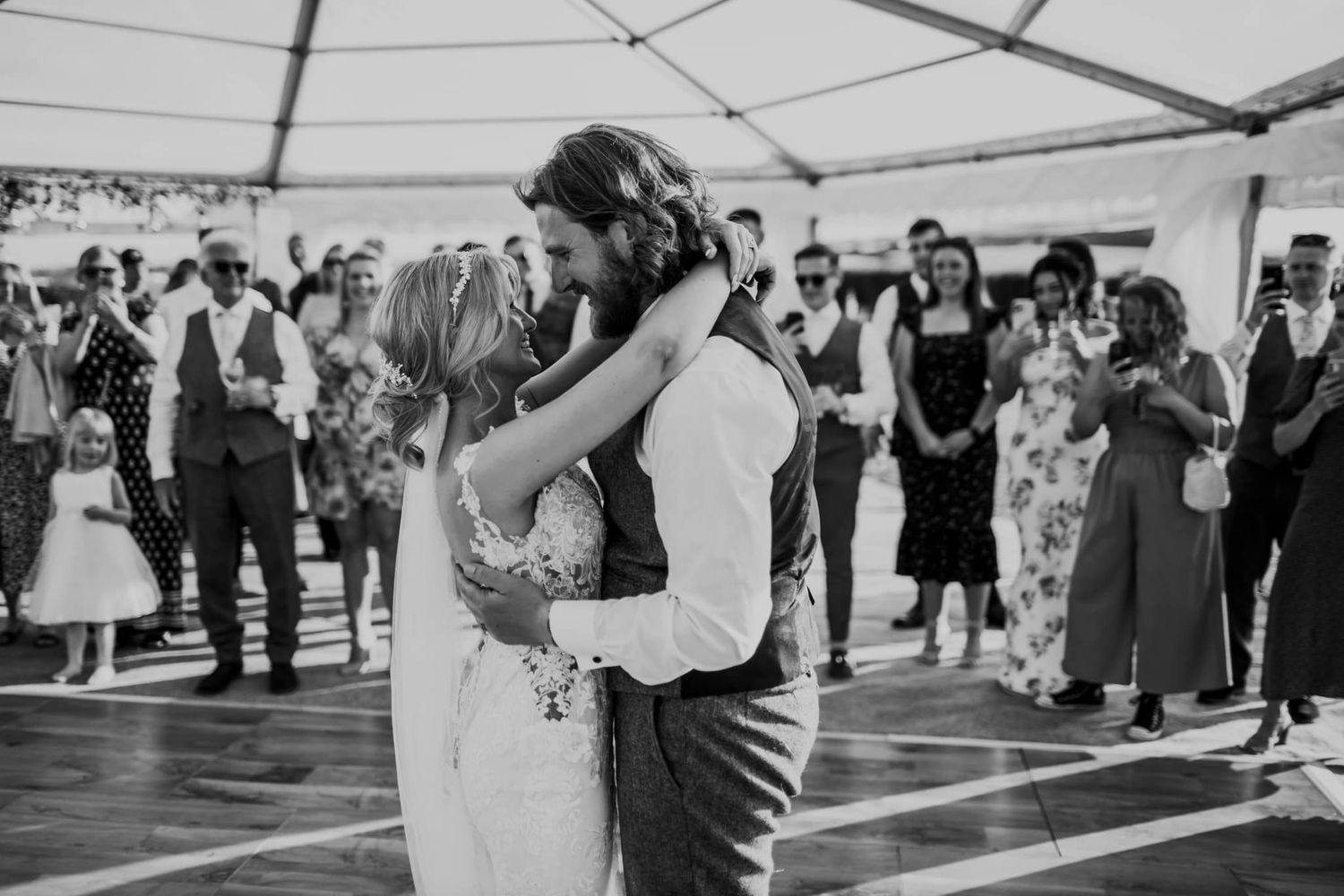 Couple shares their first dance under a reception tent while guests watch in black and white.