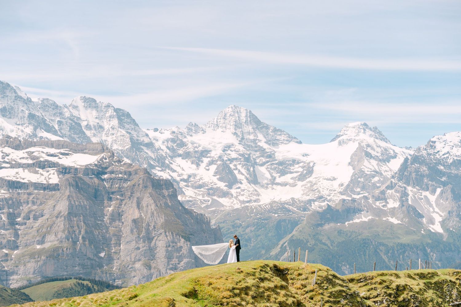 Bride and groom stand on a lush green hilltop with stunning snow-capped Swiss Alps mountains in the background.