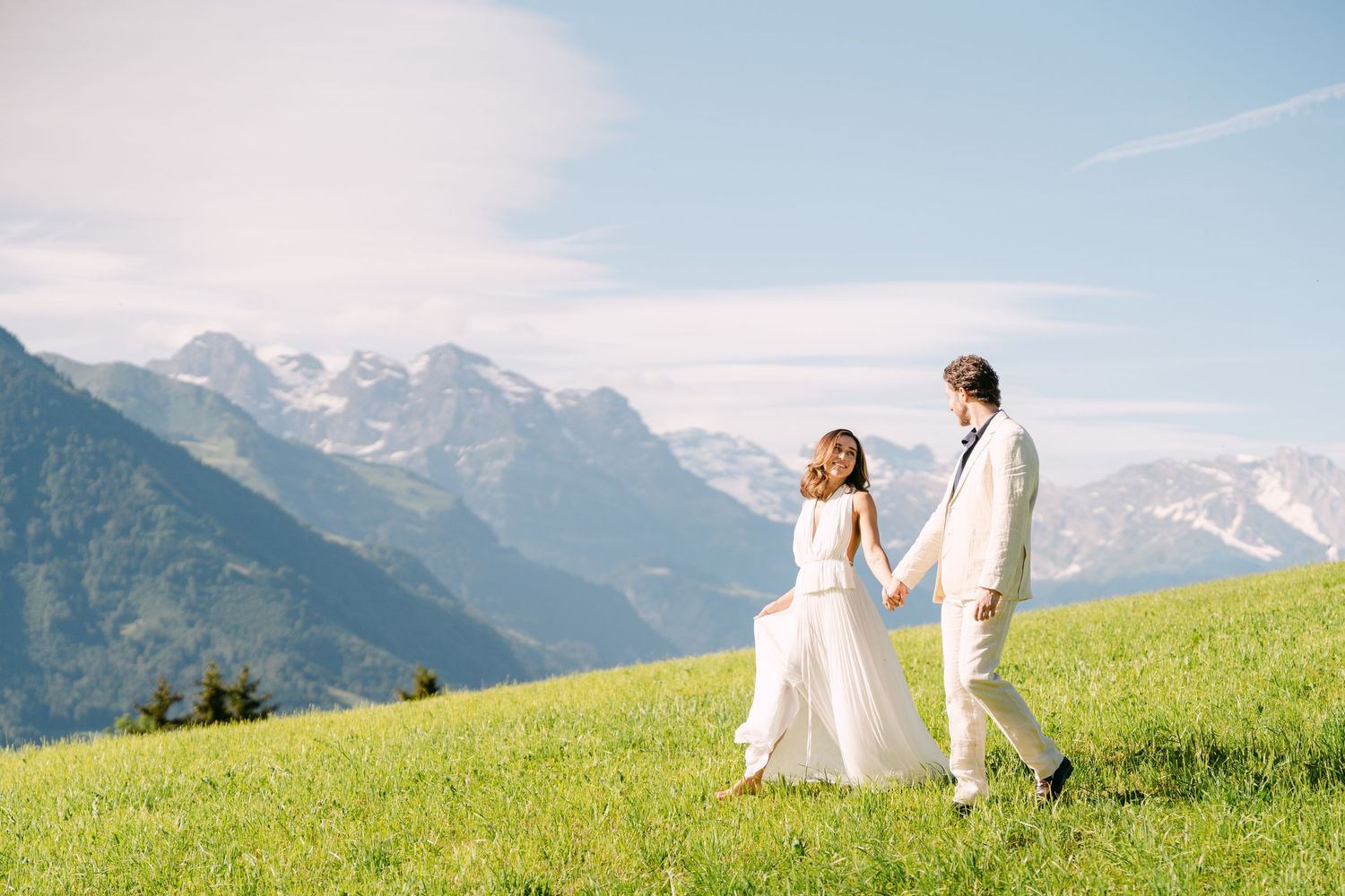 Couple in white wedding attire holding hands on a lush green alpine meadow with snow-capped mountains in the background.