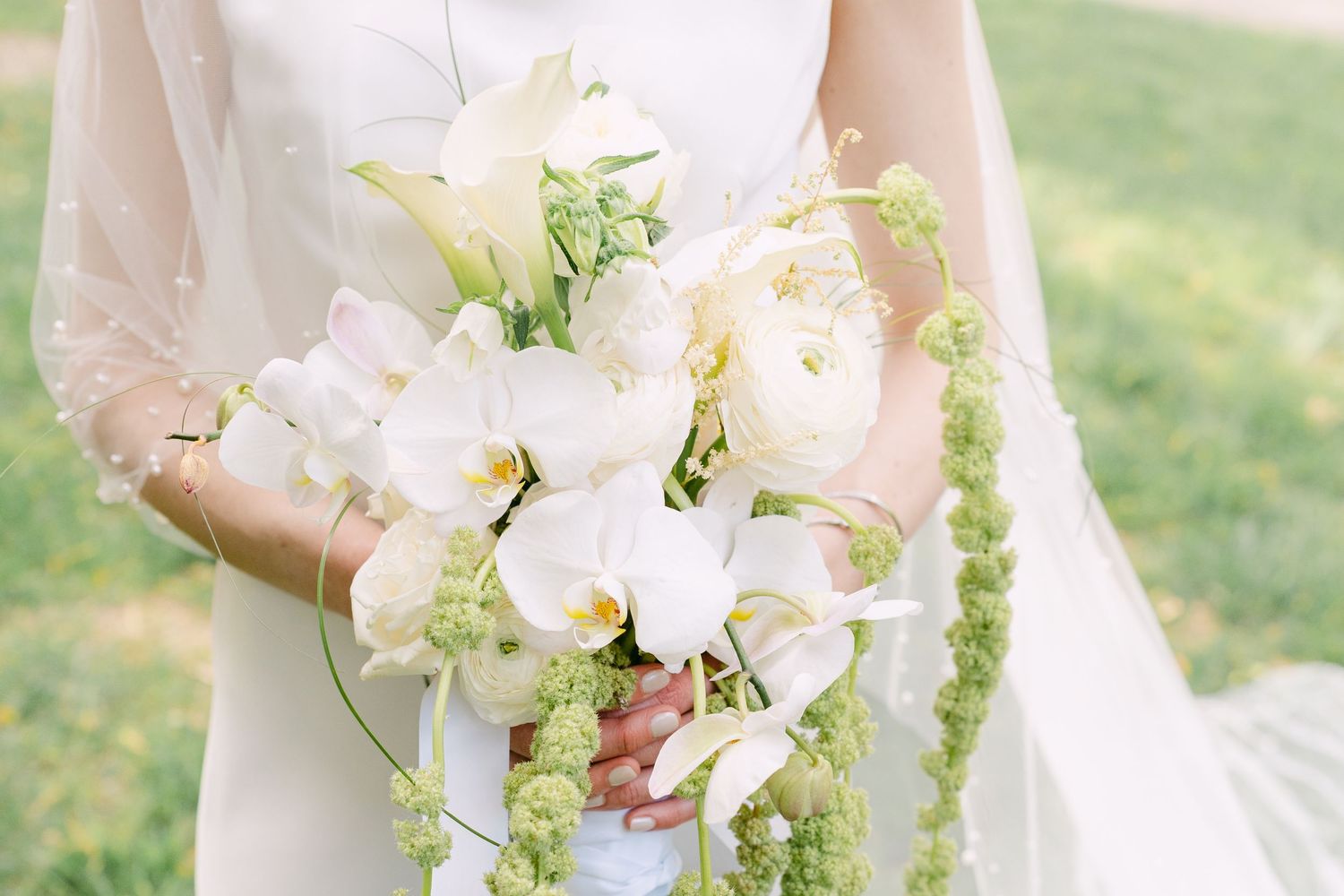 A delicate white bridal bouquet featuring lilies, roses and cascading green amaranthus against a soft outdoor background.