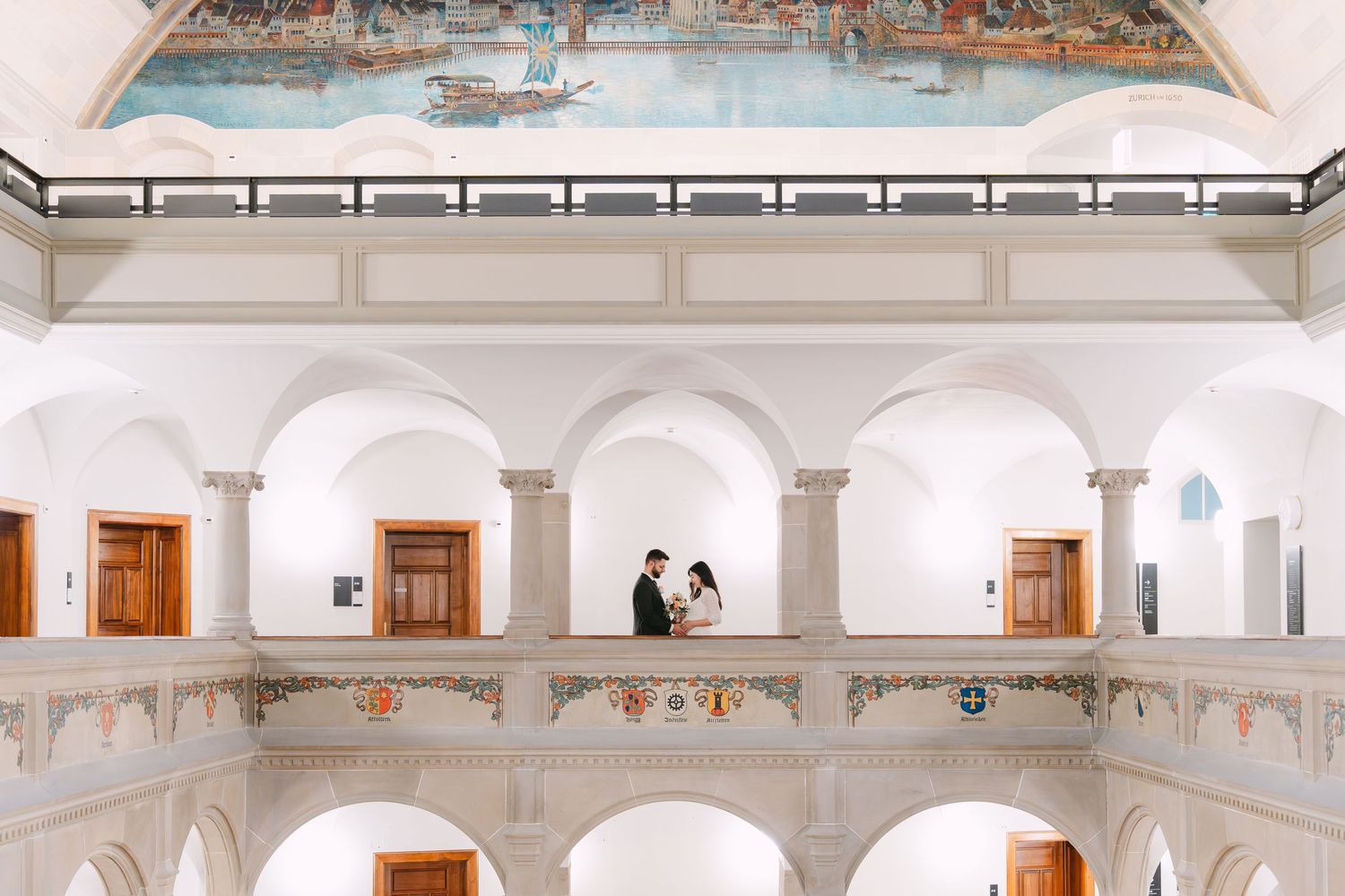 A couple stands in an elegant white archway balcony of a historic building with ornate architectural details.