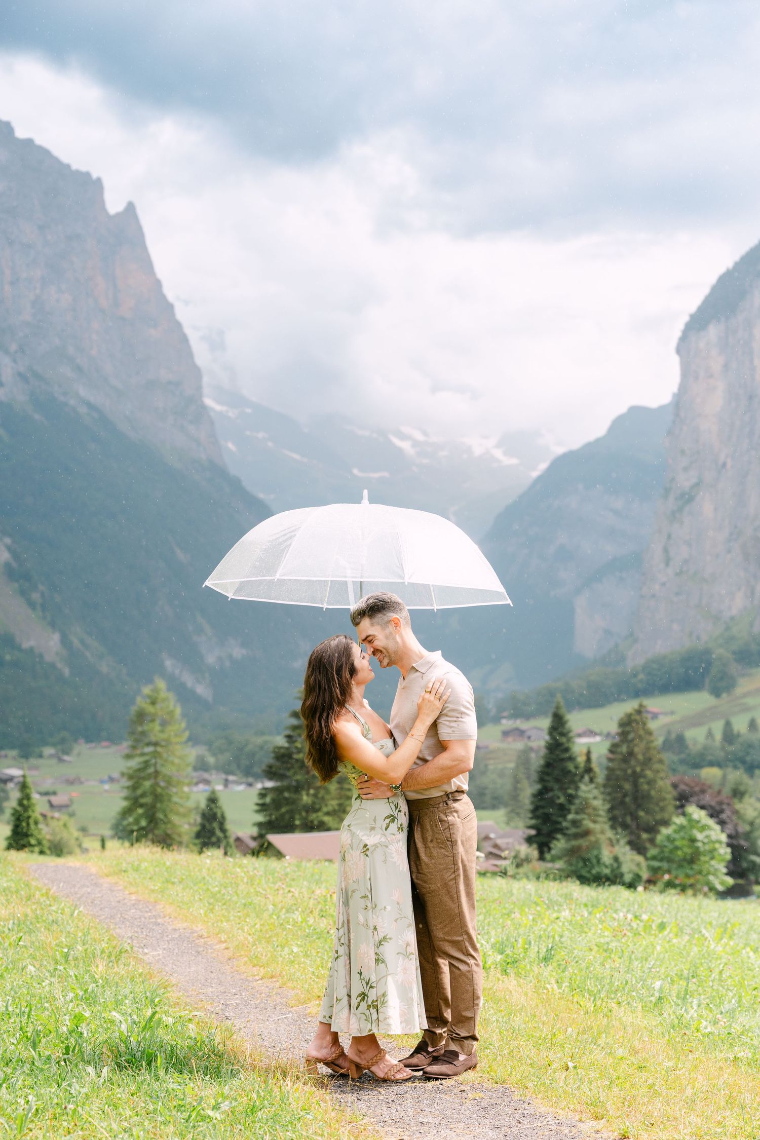 Couple embracing under clear umbrella in Swiss Alps valley with dramatic mountain backdrop and lush green meadows.