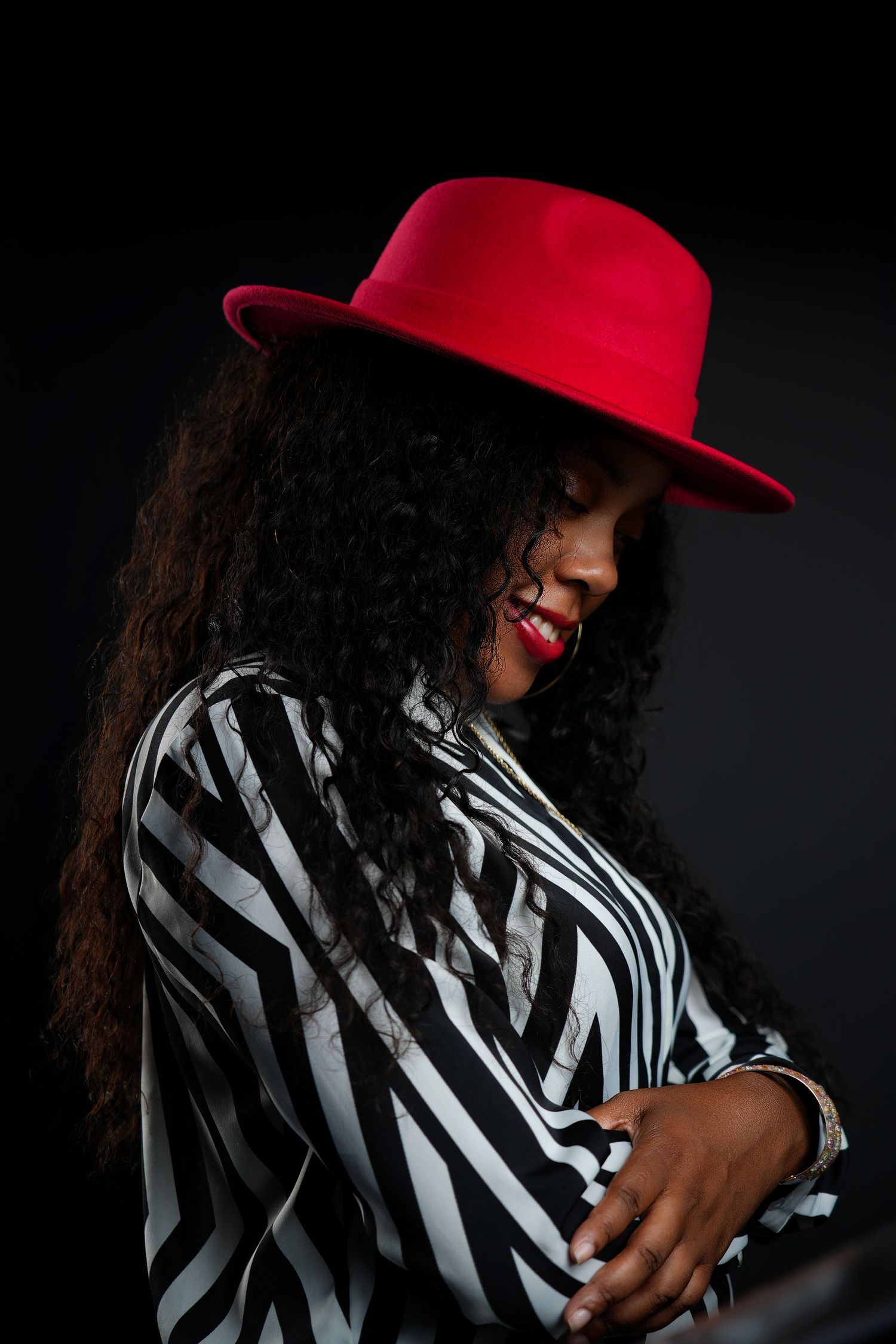 Dramatic portrait featuring red fedora and black and white striped blouse against dark background.