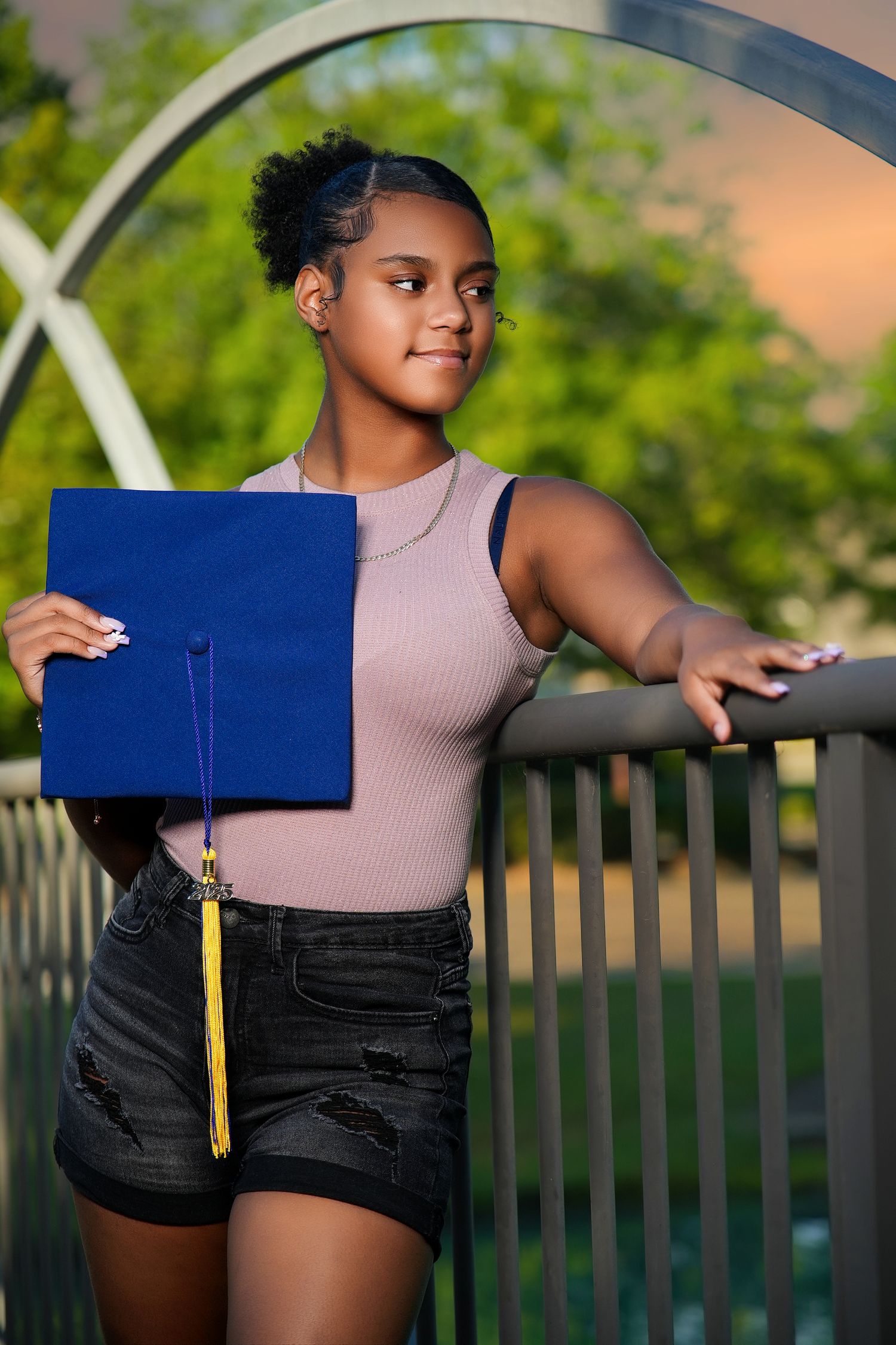 A graduate holding a blue diploma cover poses outdoors by a metal railing at sunset.