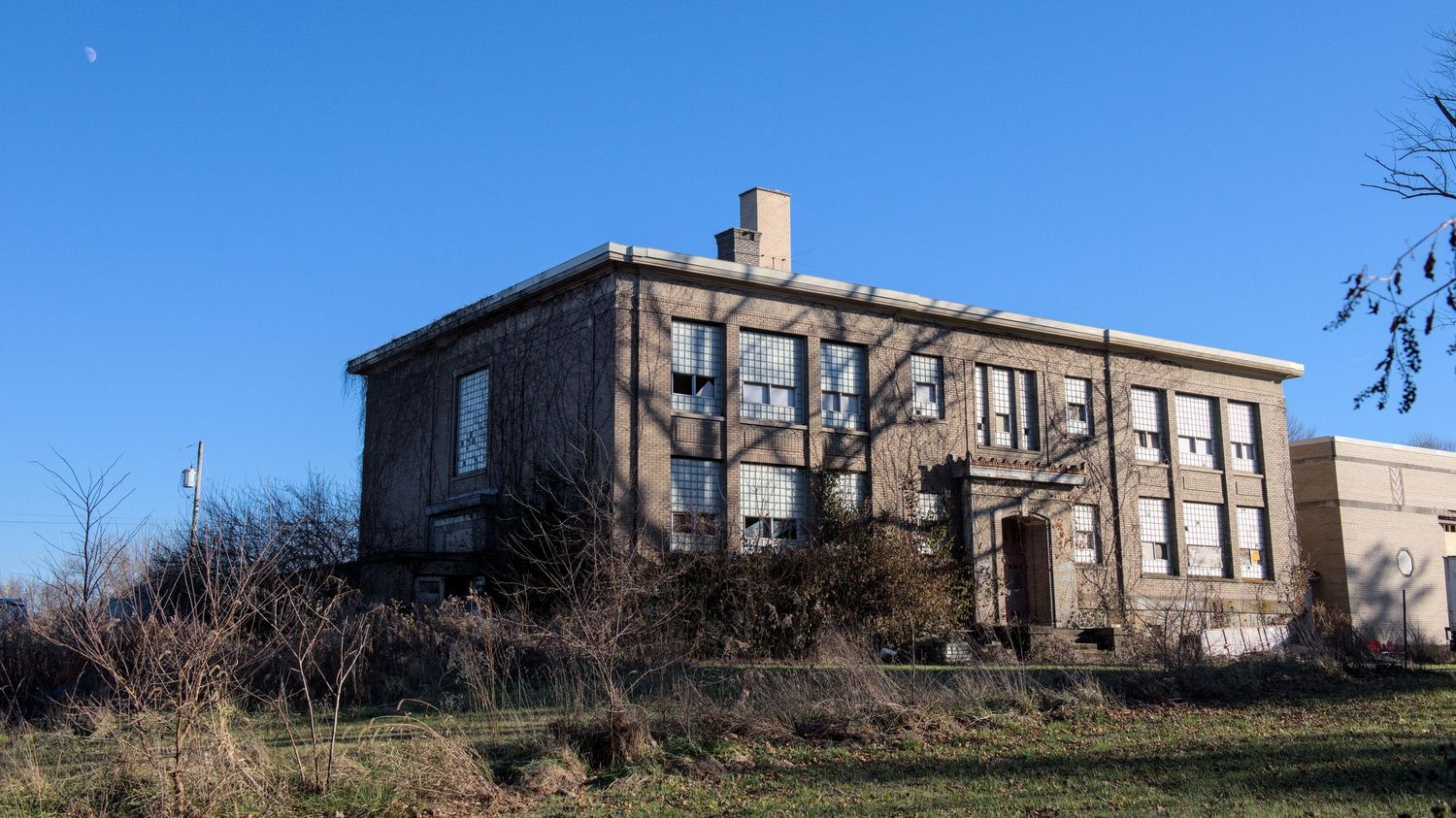 An abandoned brick school building sits covered in long shadows of a tree. A slice of moon is in the blue sky.