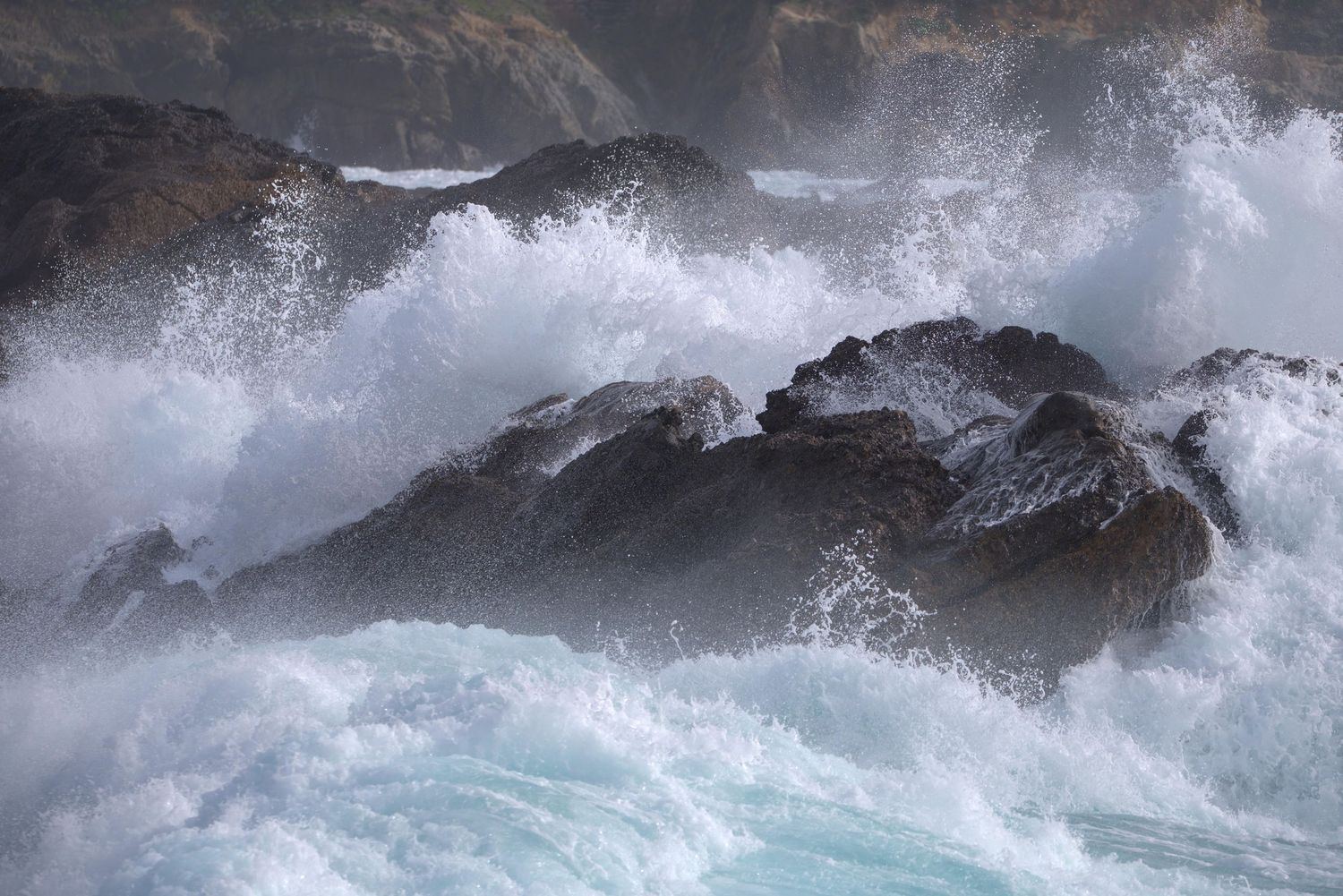 Foamy white sea spray explodes against a brown-black boulder on the coast at Point Lobos in California.