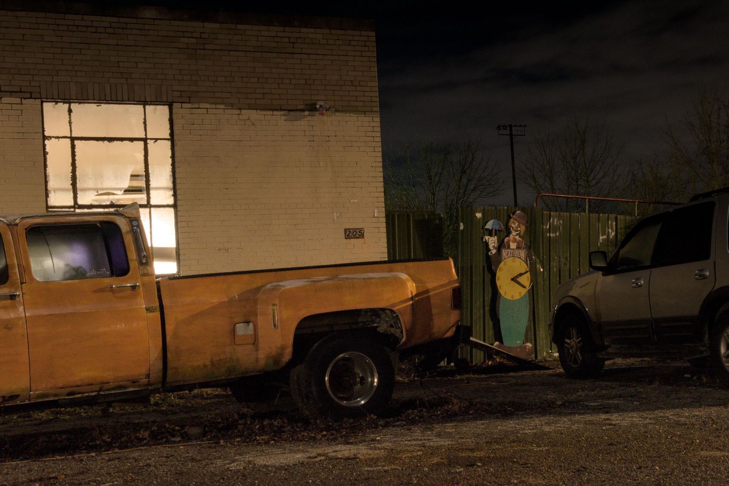 A night time image showing an old yellow truck beside a brick building with a broken window and a clown carnival sign.
