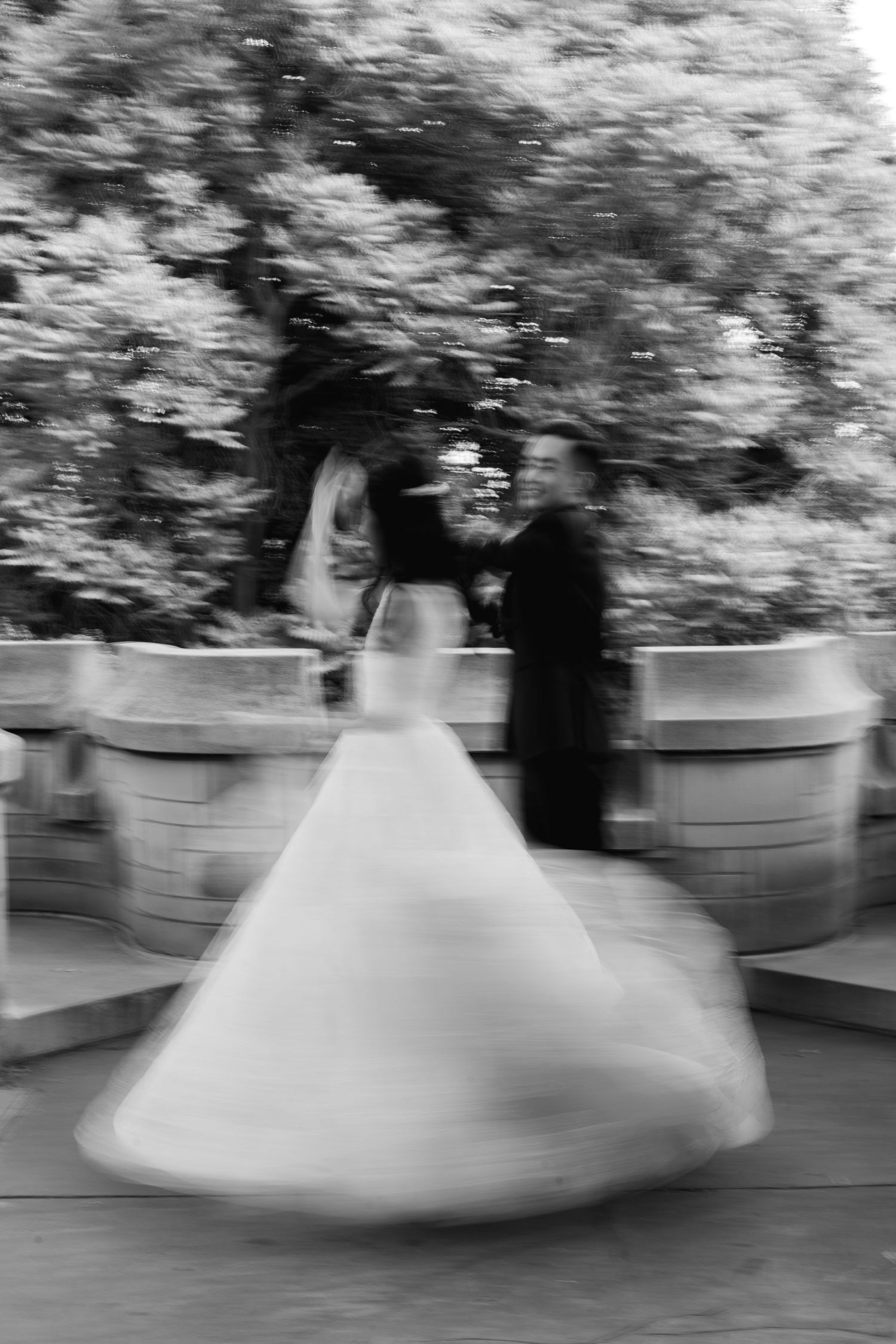 Black-and-white wedding portrait of a bride twirling in her flowing gown beside the groom, with soft motion blur and trees in the background