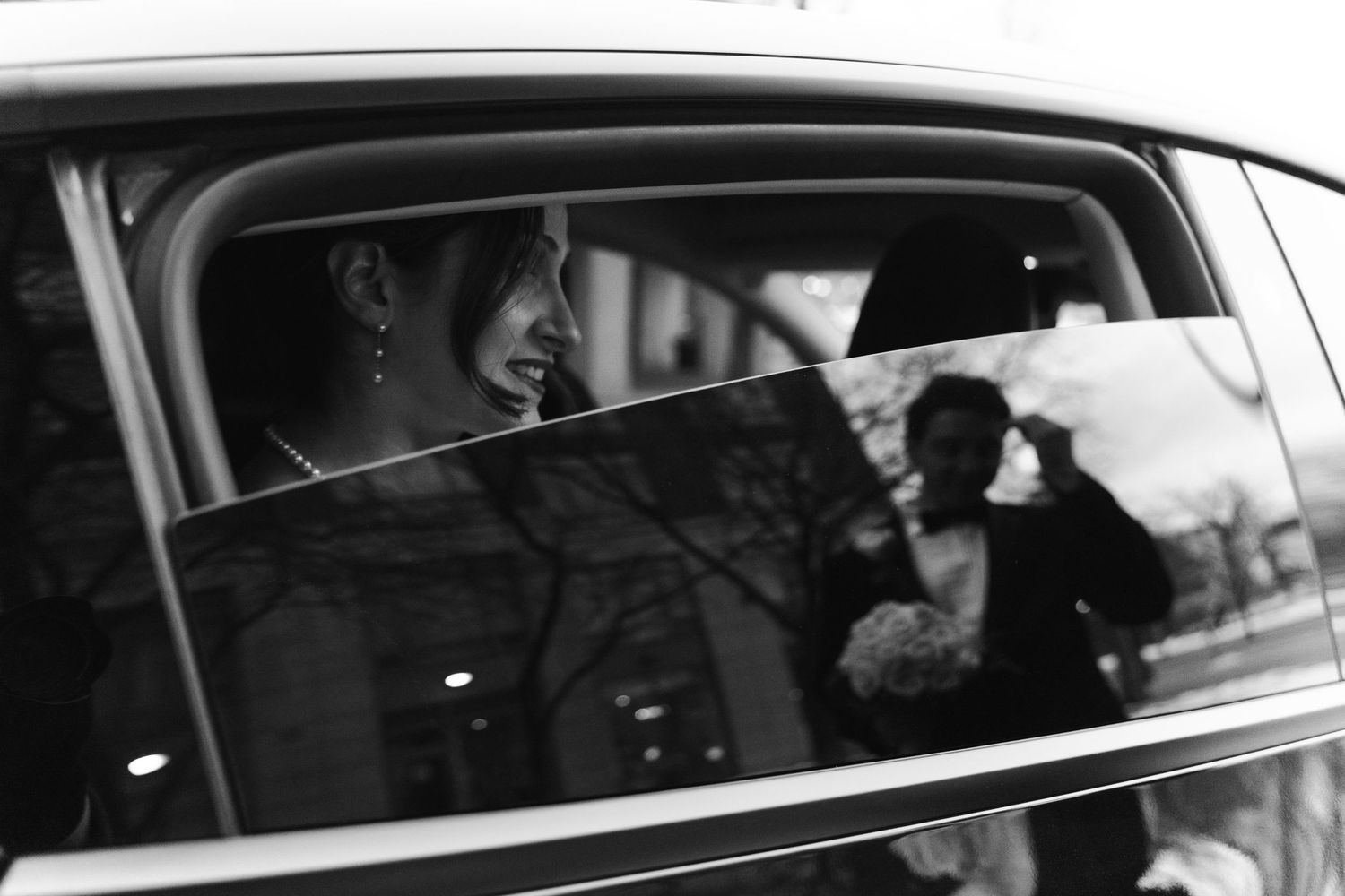 Black-and-white candid photograph of a bride smiling inside a car, with the groom reflected in the window beside her.