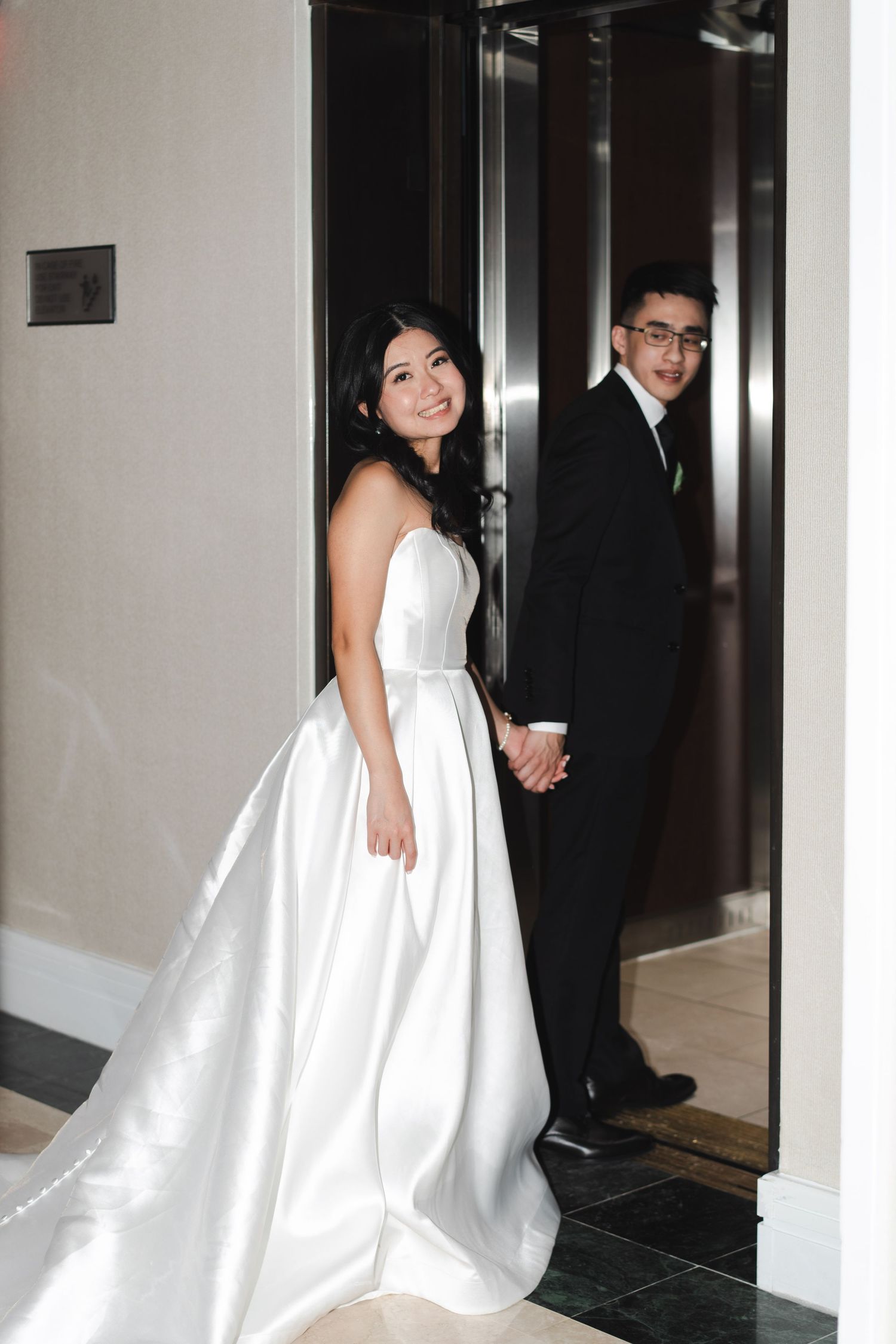 Bride in a flowing white wedding gown looking back toward the camera while the groom stands beside her in a hotel hallway.