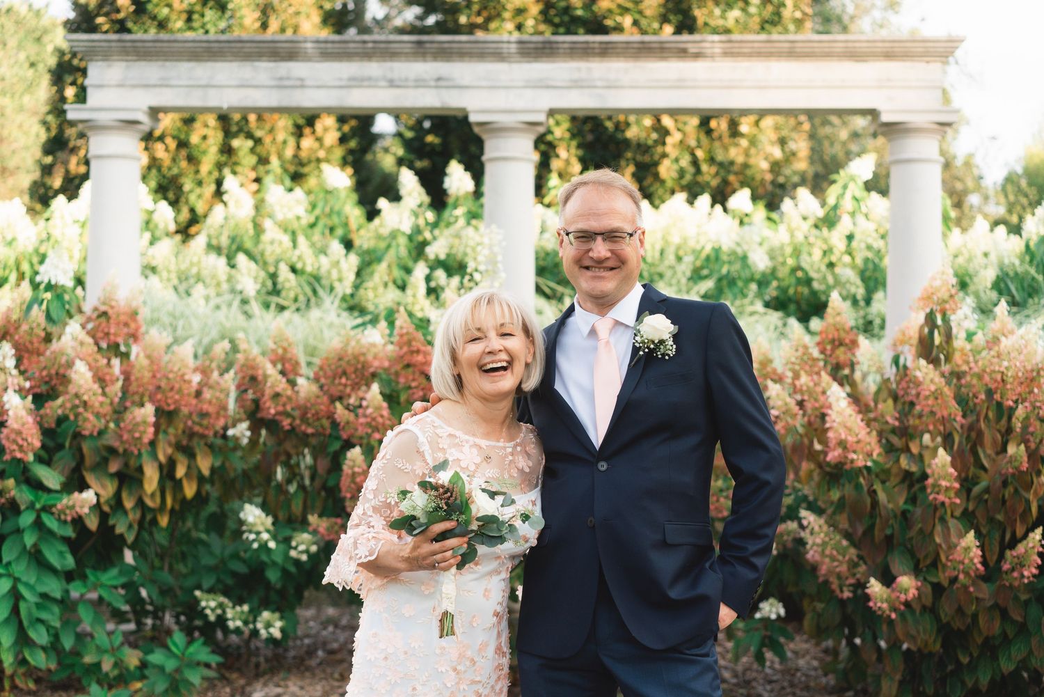 Newlyweds Jolanta and Rick smile together beneath a garden pergola surrounded by colourful flowers and greenery in Oshawa.