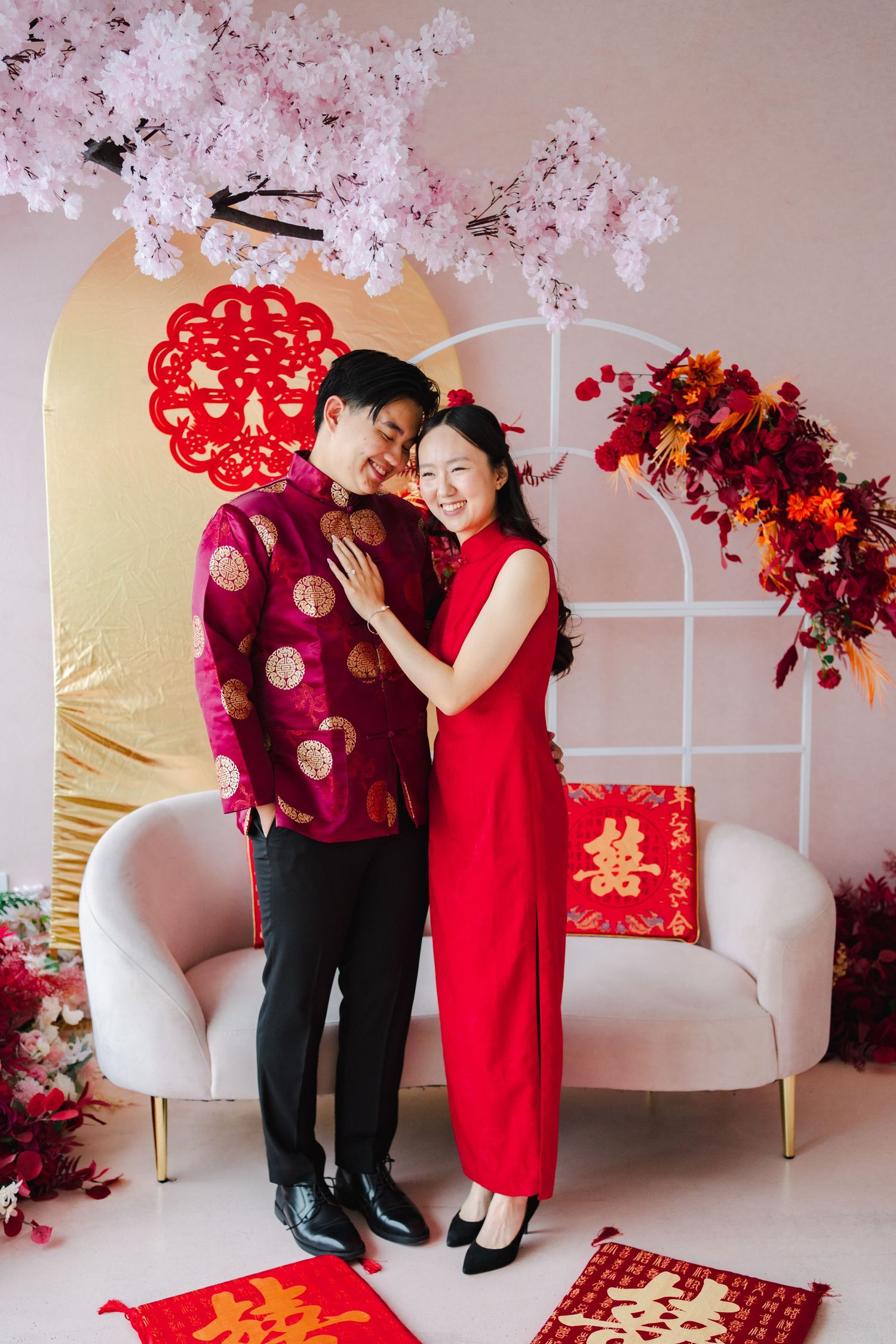 Couple in traditional red Chinese wedding attire embracing beneath pink floral decorations at ESpace Studios.