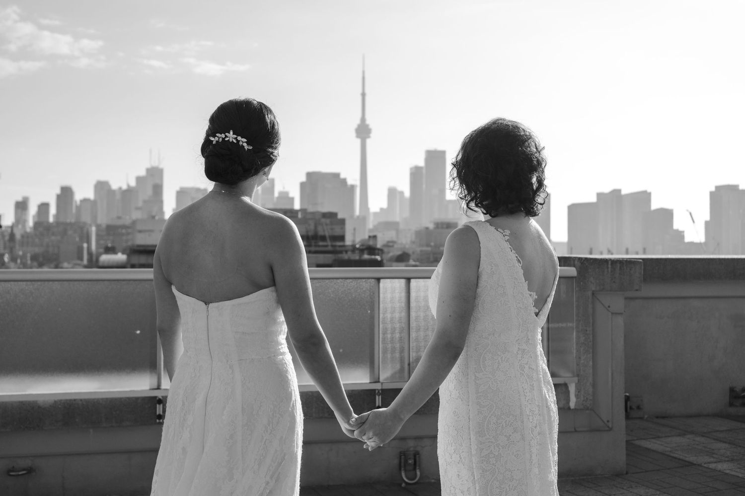 Two brides holding hands on a rooftop terrace overlooking the Toronto skyline and CN Tower in a black-and-white wedding photograph.