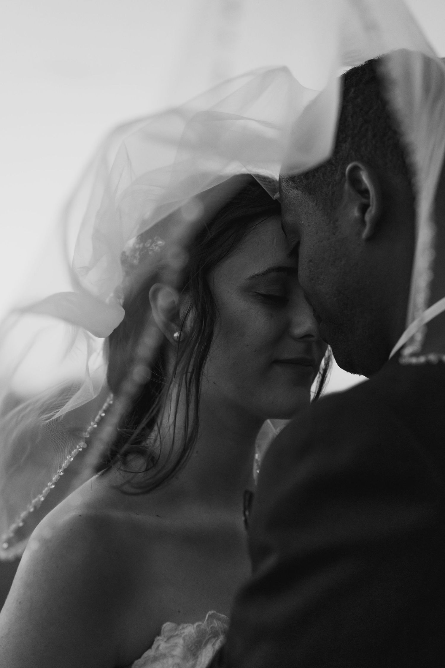 Intimate black-and-white wedding portrait of a bride beneath her veil, leaning close to the groom with their foreheads nearly touching.