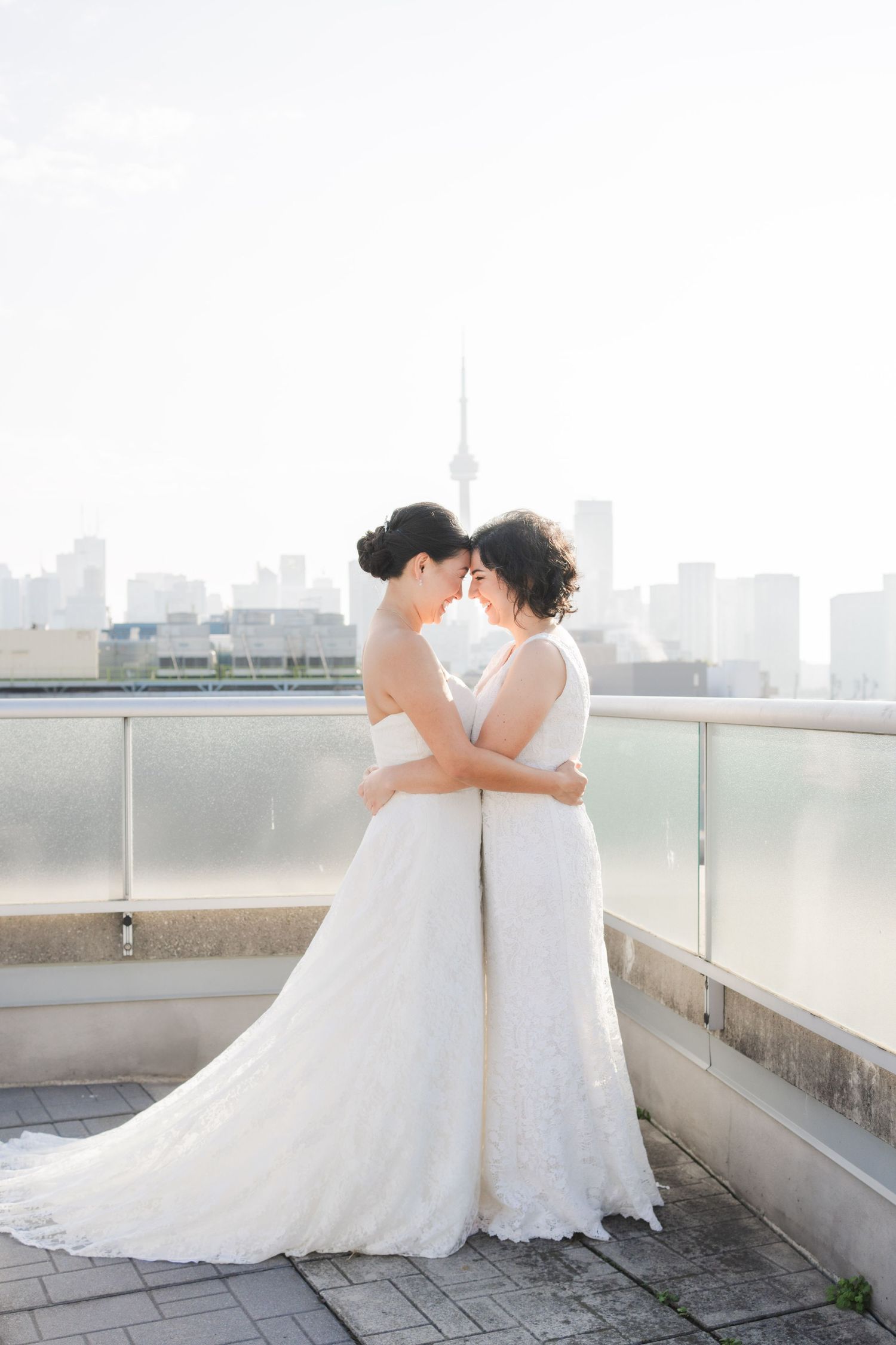 Two brides embracing on a rooftop terrace overlooking the Toronto skyline, with their white wedding gowns flowing across the ground.