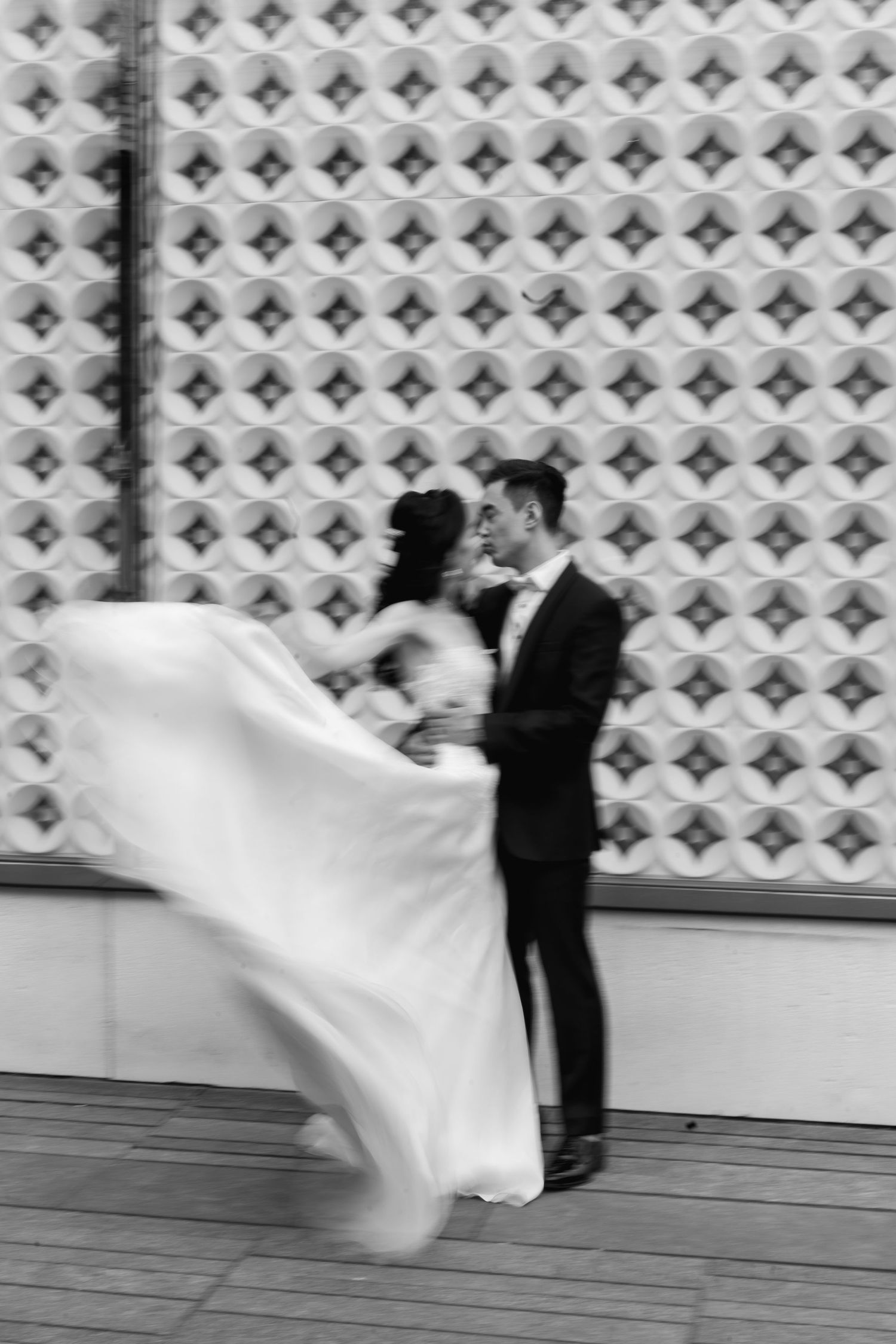 Black-and-white wedding photograph of a groom watching the bride's flowing dress sweep through the air against a patterned architectural wal
