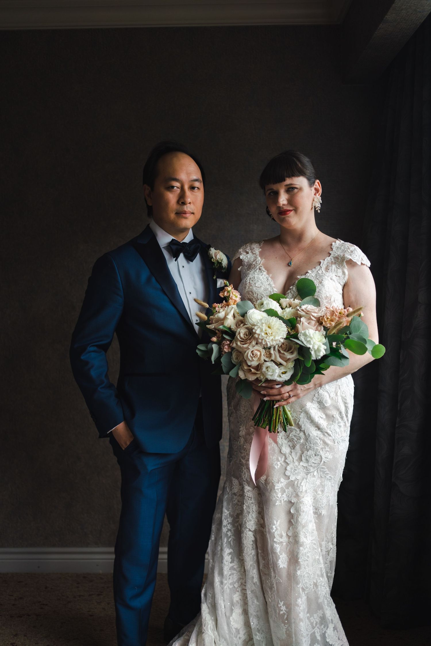 Bride holding her wedding bouquet beside the groom in a blue suit, captured in a dramatic low-light wedding portrait.
