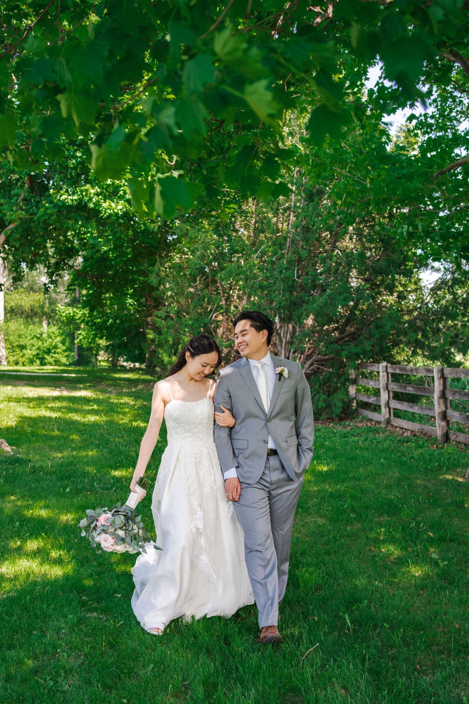 Bride and groom walking together through a lush green garden beneath leafy trees on their wedding day.