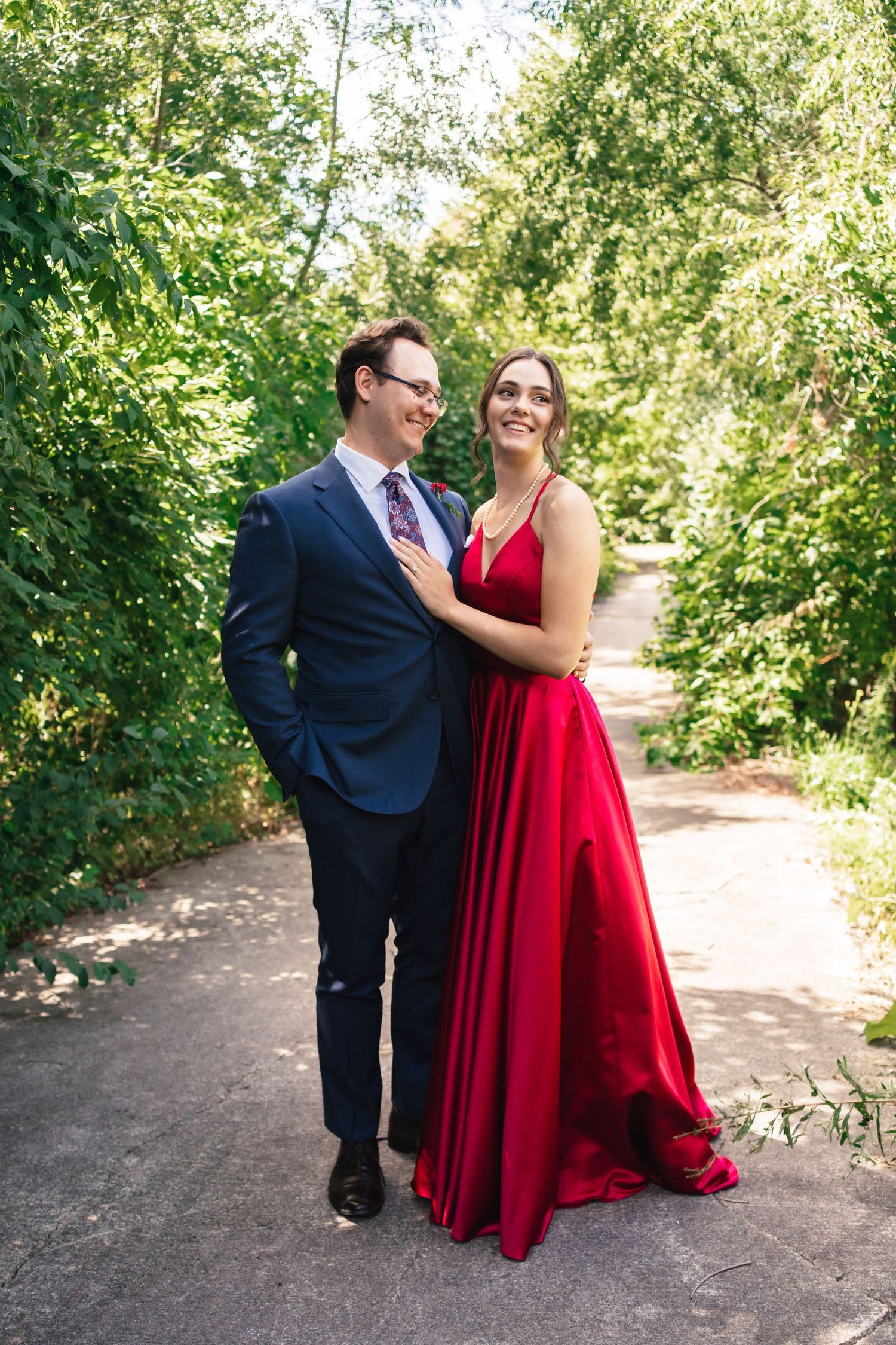 Bride in a dramatic red wedding gown standing with her groom in a navy suit on a tree-lined path during their intimate backyard wedding.