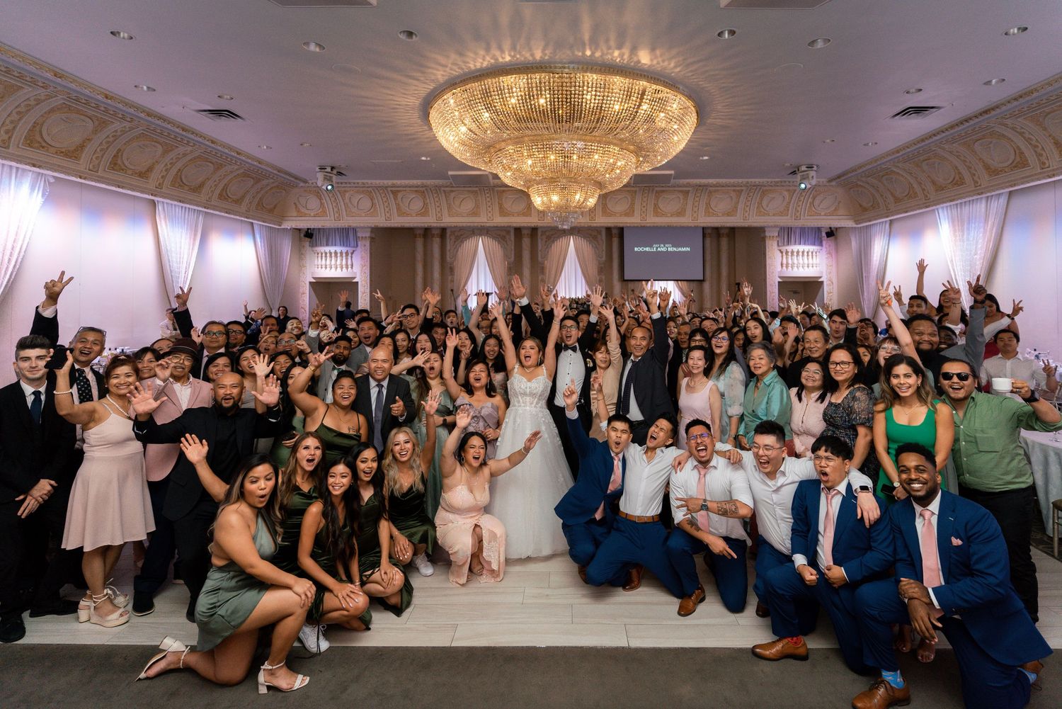 Large group of wedding guests celebrating together in an elegant ballroom beneath a chandelier.