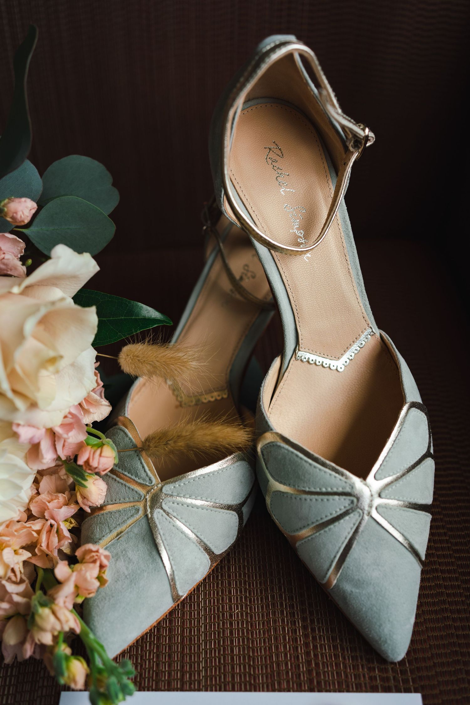 Elegant bridal shoes with delicate ankle straps arranged beside a pastel wedding bouquet on a dark wooden surface.