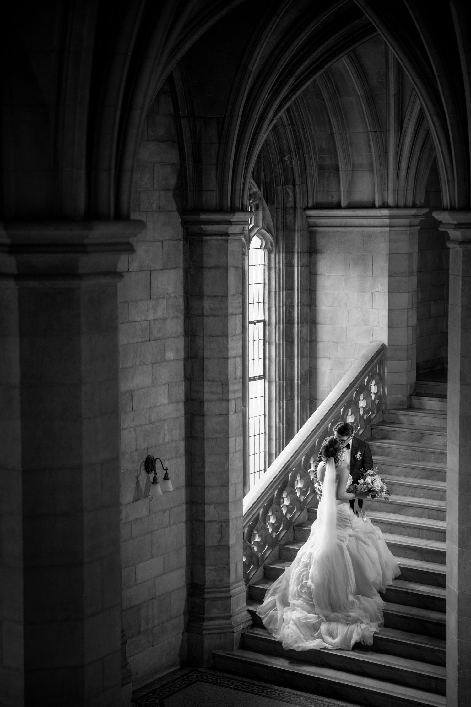 Black-and-white bridal portrait on a grand stone staircase framed by Gothic arches and tall windows at Knox College in Toronto.