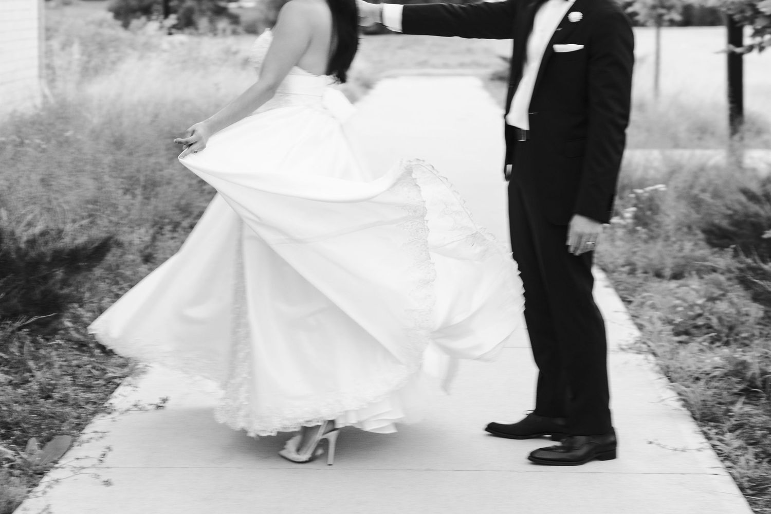 Black-and-white wedding photograph of a bride lifting her flowing gown while walking beside the groom on an outdoor path.