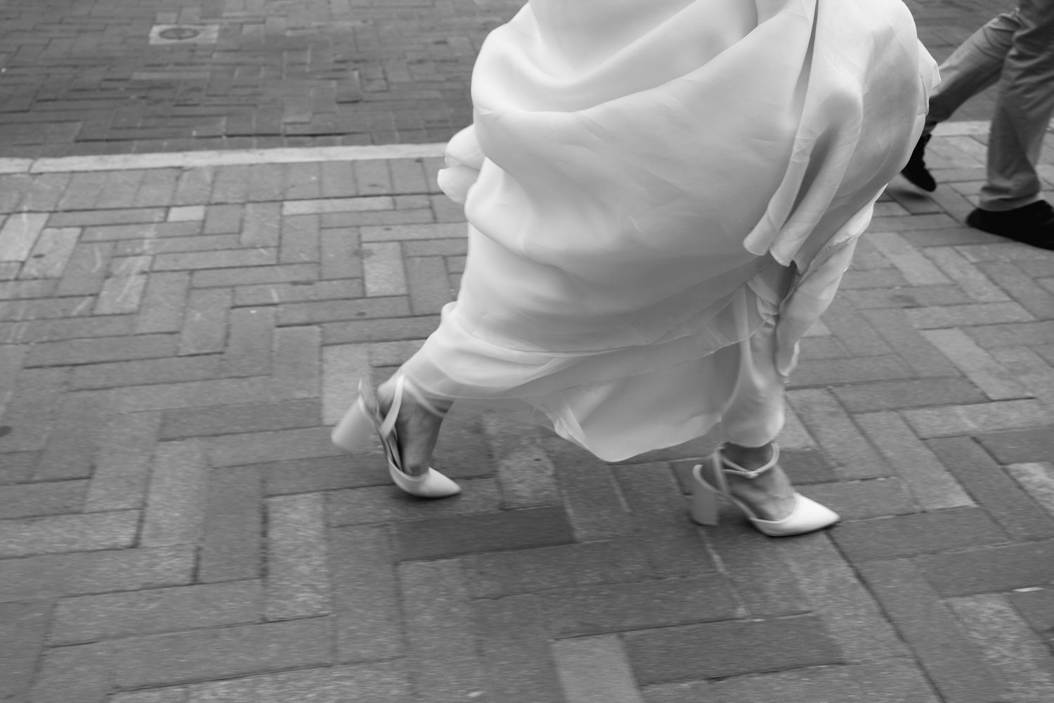 Black-and-white detail photograph of a bride lifting her wedding dress while walking in elegant heels along a brick-paved sidewalk.