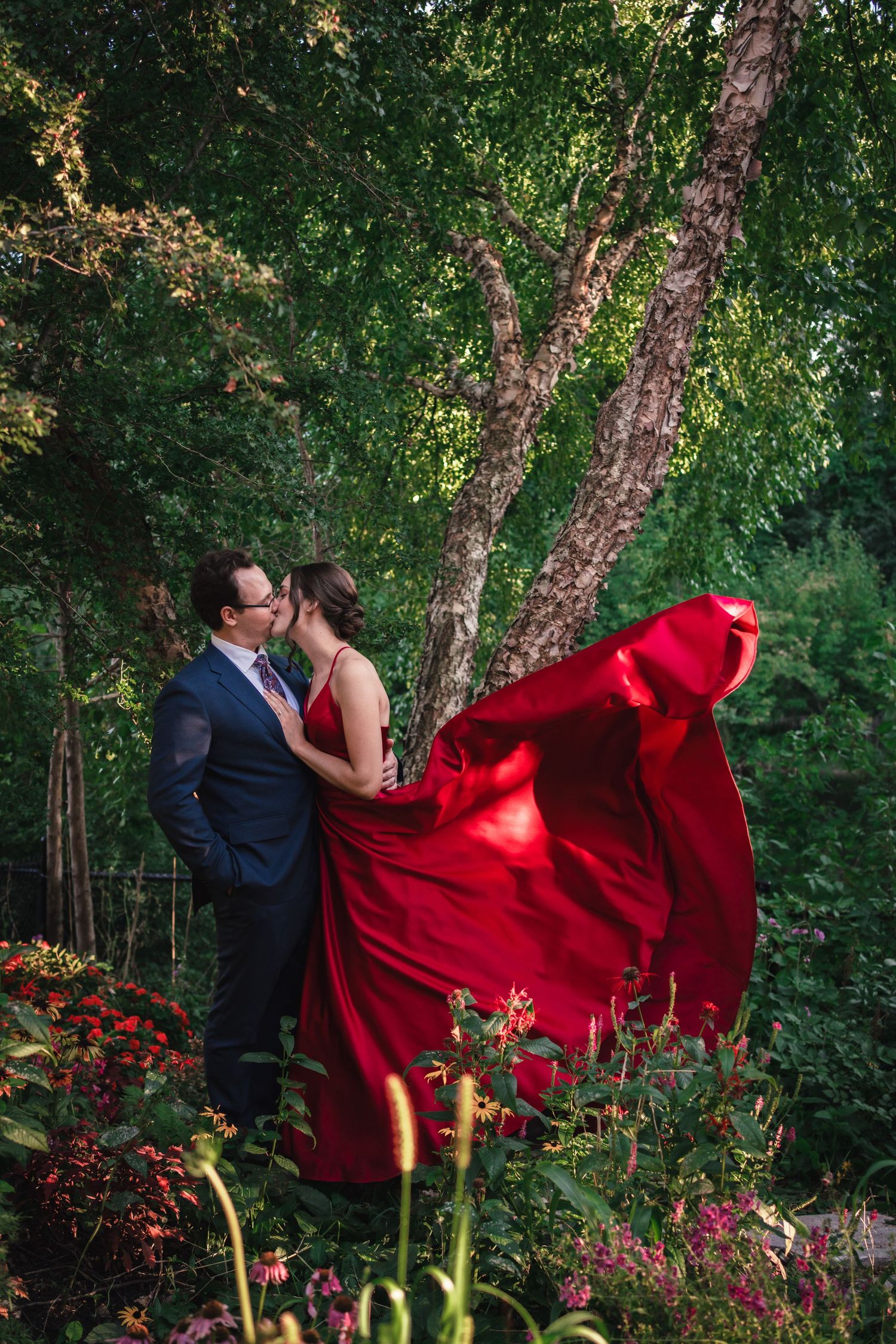 Groom lifting the bride in her dramatic flowing red wedding gown amid lush greenery and colourful flowers during their intimate backyard wed