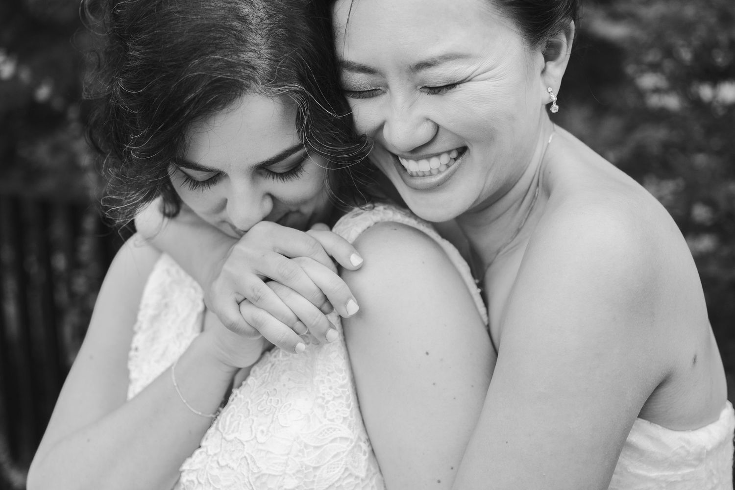 Black-and-white portrait of two brides sharing a tender embrace, one kissing the other's hand while her partner smiles.