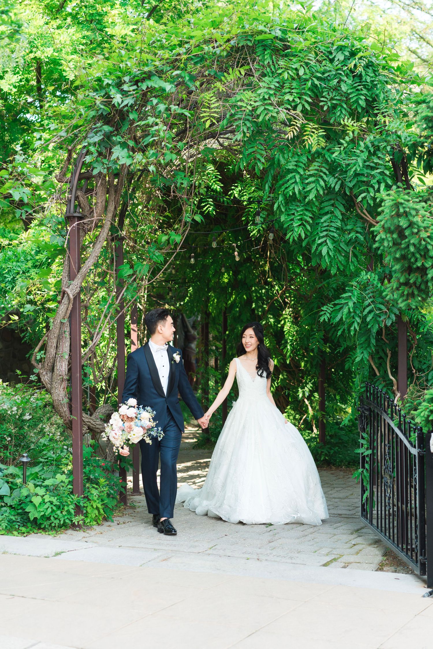 Bride and groom standing together beneath a lush green archway of hanging vines during their outdoor wedding portraits.