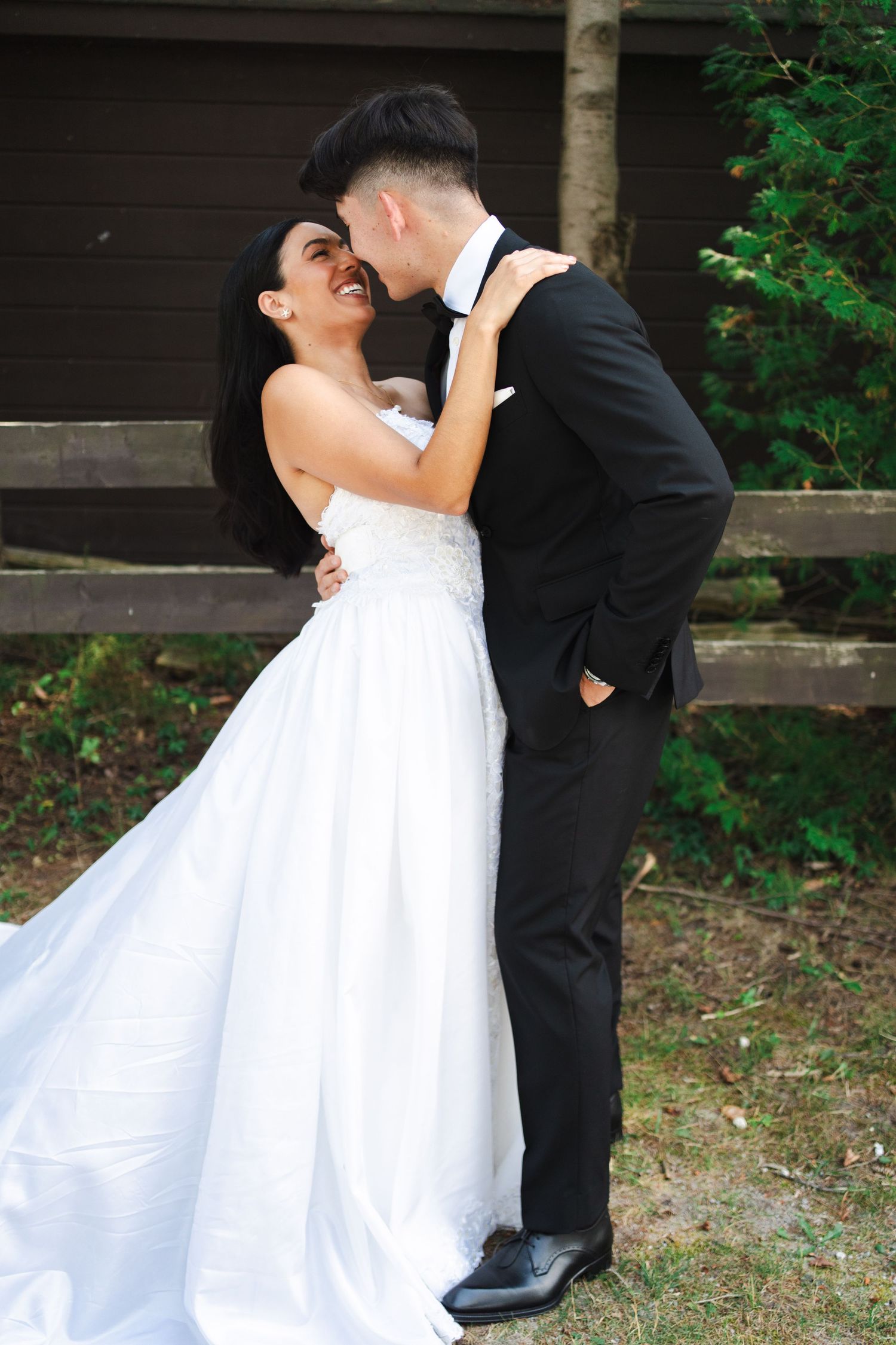 Bride and groom sharing an intimate kiss outdoors beside a rustic wooden building, surrounded by greenery.