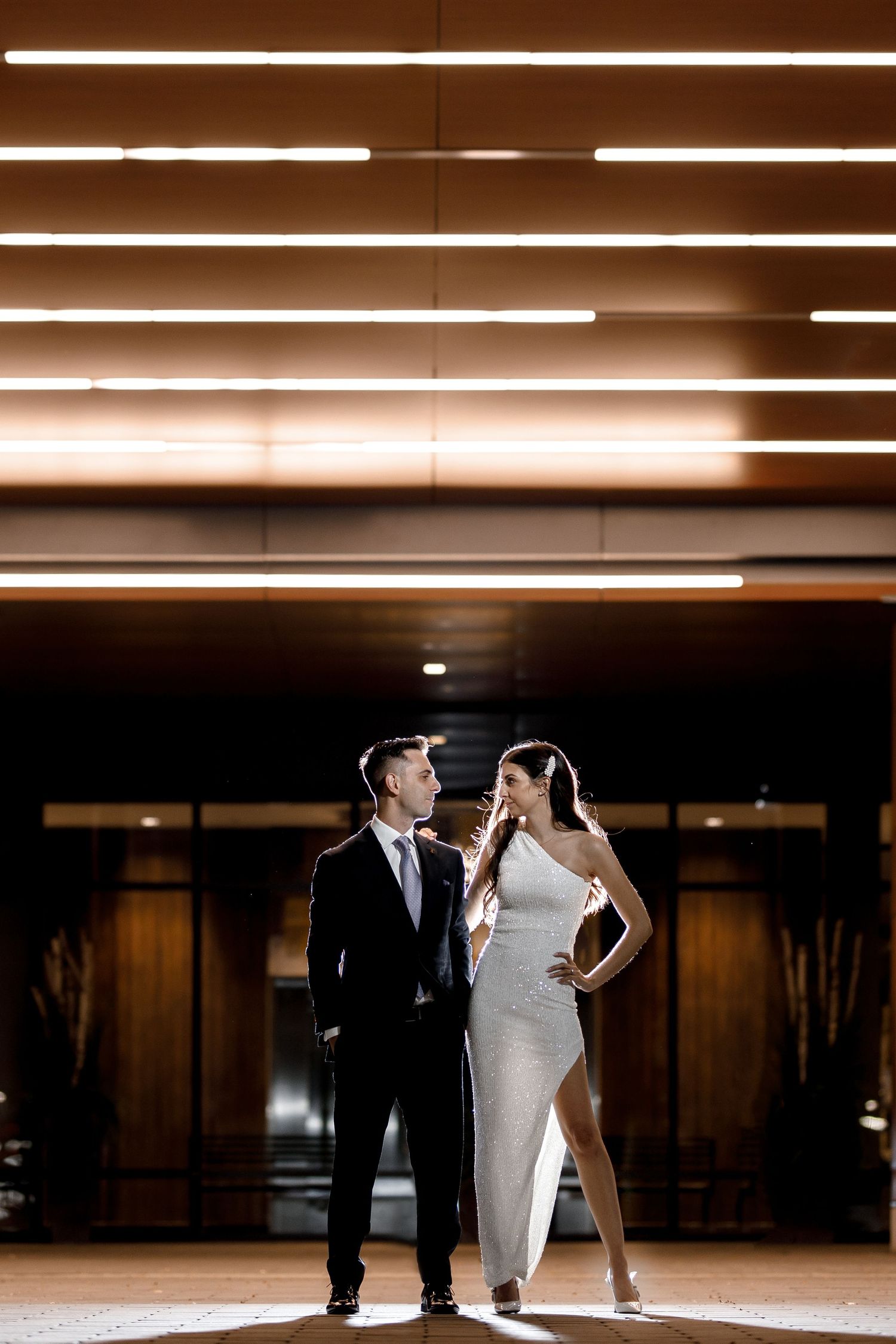 Bride and groom posing together beneath warm horizontal ceiling lights in a modern wedding reception venue.