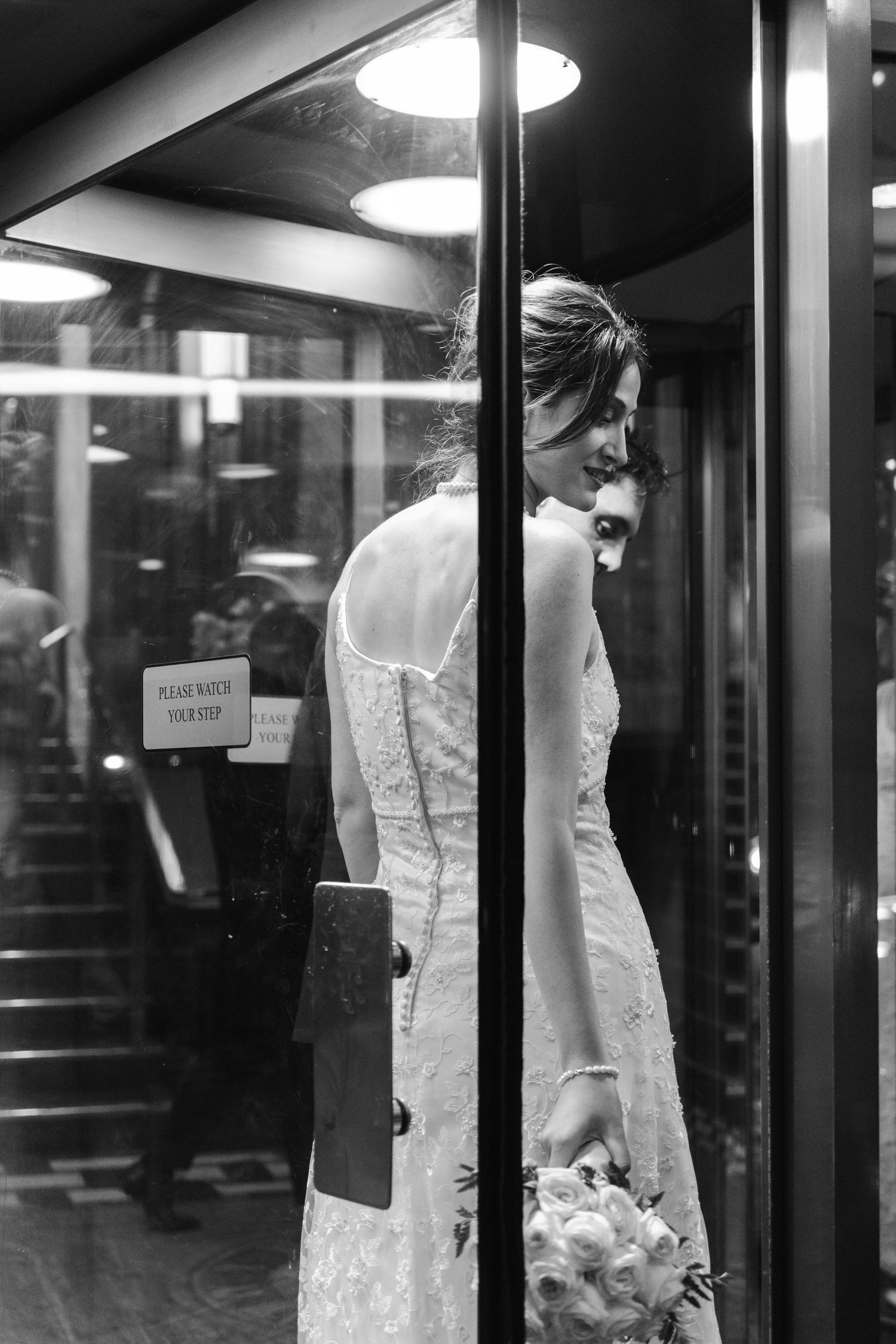 Black-and-white portrait of a bride in her wedding gown standing inside a glass elevator with reflections framing the scene.