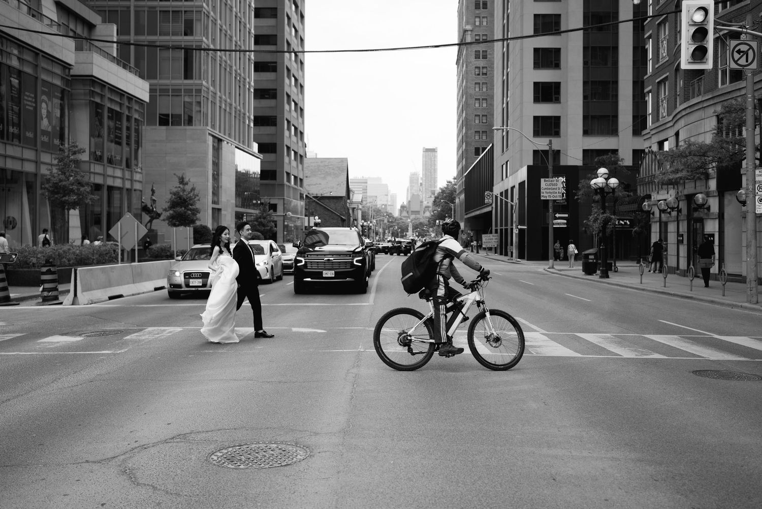 Black-and-white street photograph of a bride and groom crossing a busy downtown Toronto intersection as a cyclist passes in the foreground.