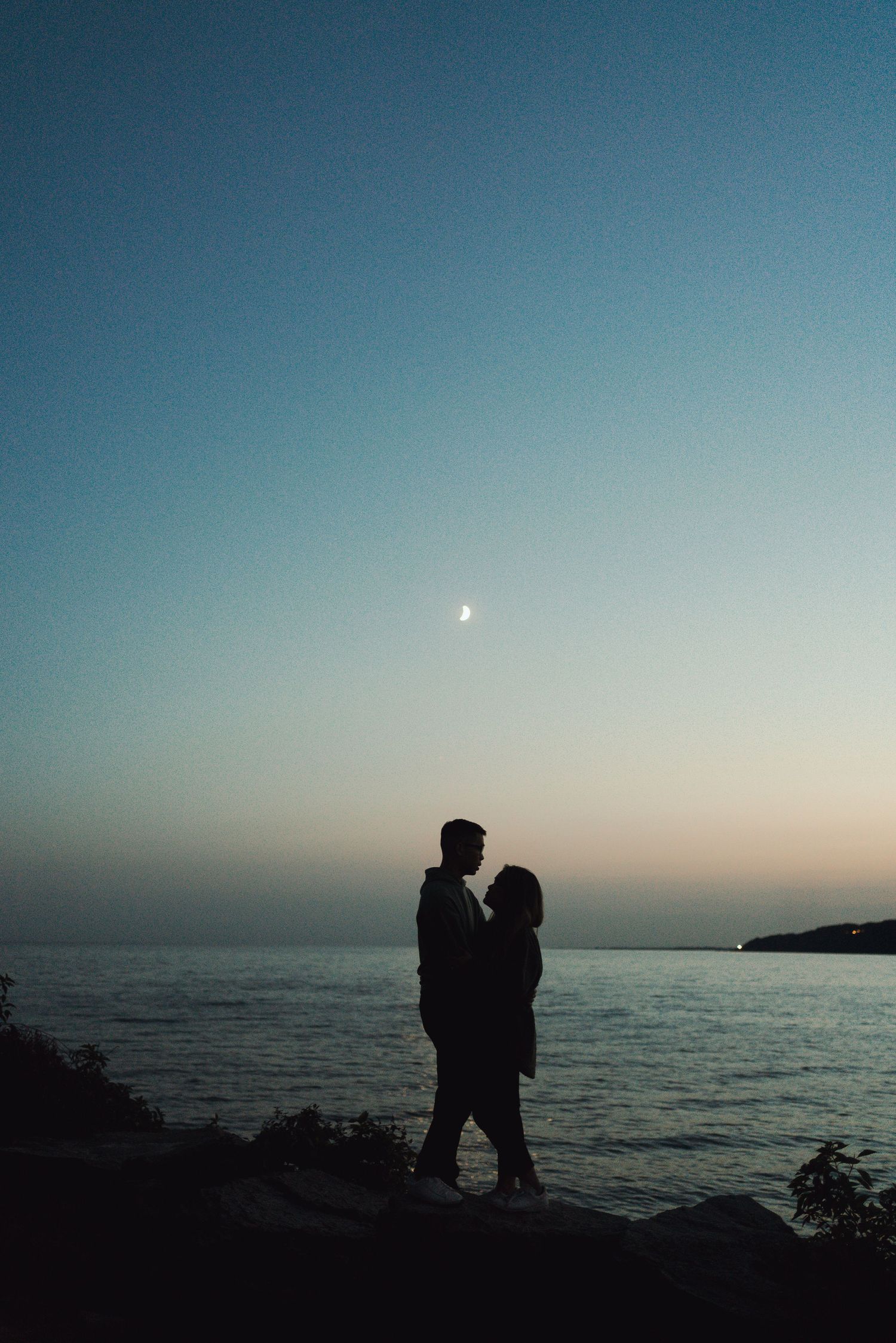 Silhouette of a couple embracing beside the water at dusk, beneath a glowing evening sky with a crescent moon at Scarborough Bluffs