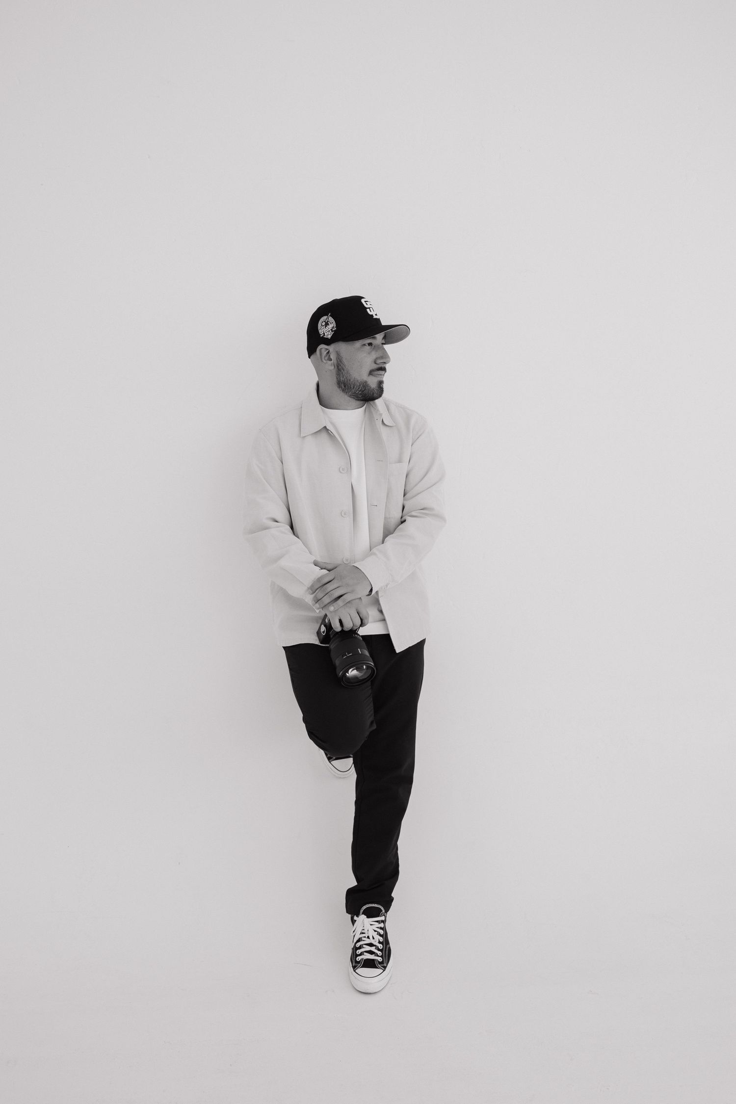 Black and white portrait of a man in a cap and light jacket holding a camera, leaning against a white wall.
