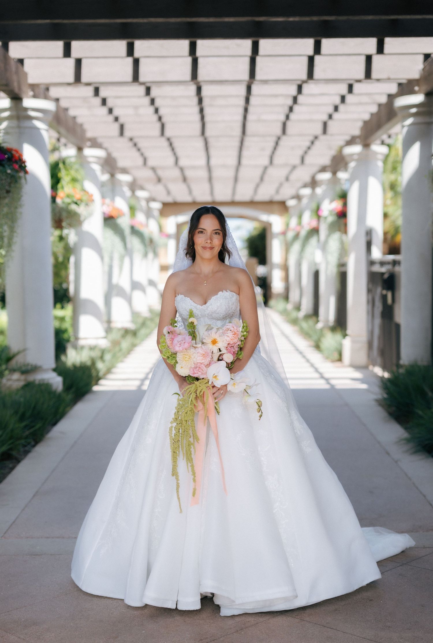 Bride in strapless ball gown wedding dress holding colorful bouquet standing under pergola in elegant outdoor venue.